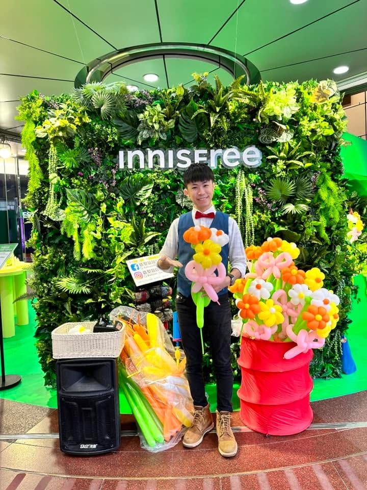 innisfree 門市綠植牆前，柯男拿著花朵氣球花束，旁邊紅桶裡插滿橘黃白花朵