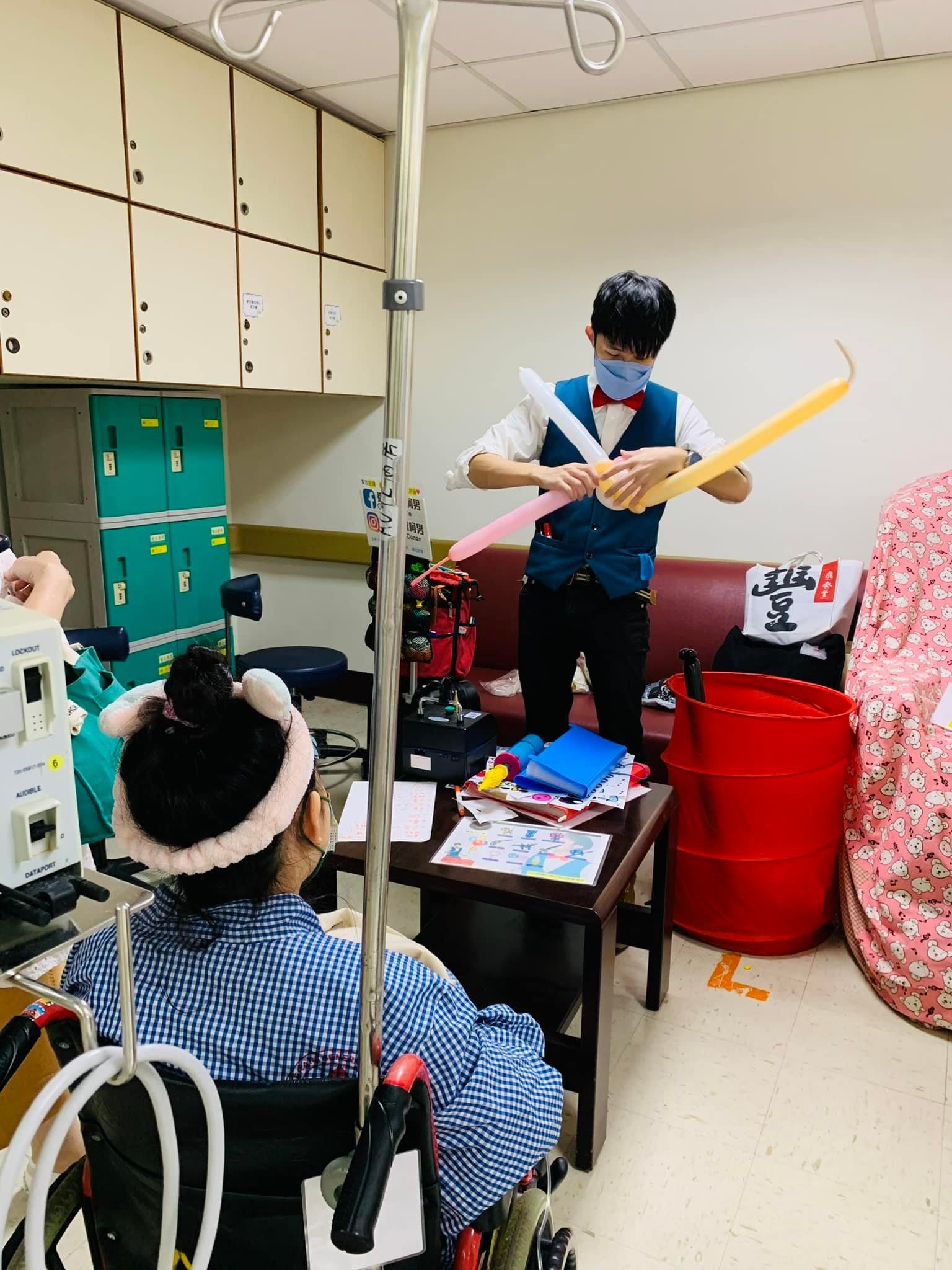 柯男拉著粉白黃長條氣球表演，坐輪椅的病童背對鏡頭在前方觀看