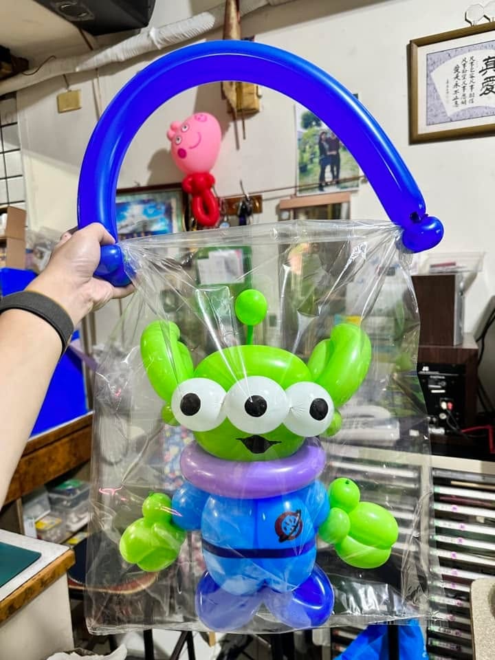 裝在透明塑膠袋裡的三眼怪造型氣球，袋口用深藍色長條氣球做成提把，一隻手提著袋子