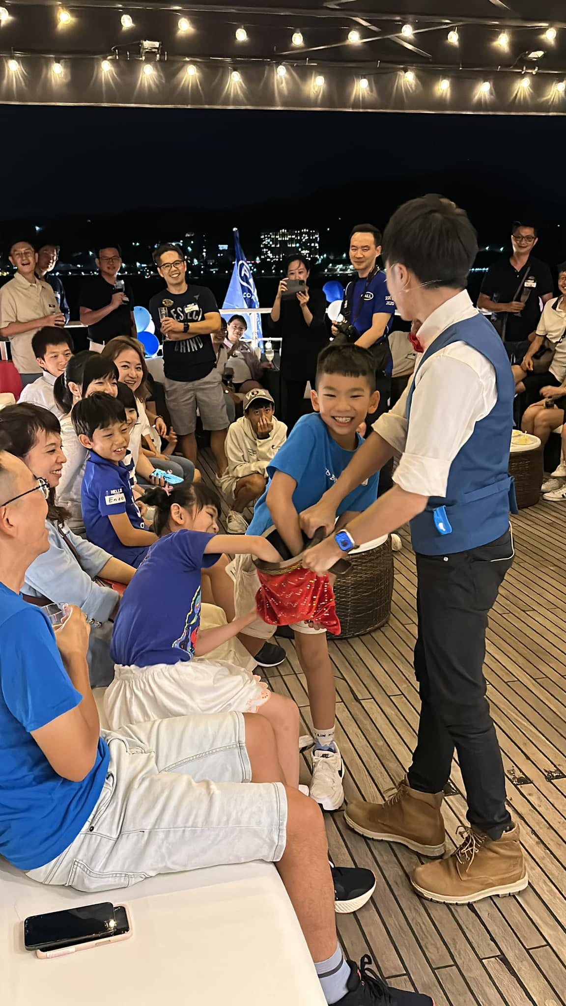 柯男請穿藍色上衣的男孩上台拉紅色魔術布，船上的大人小孩笑著看