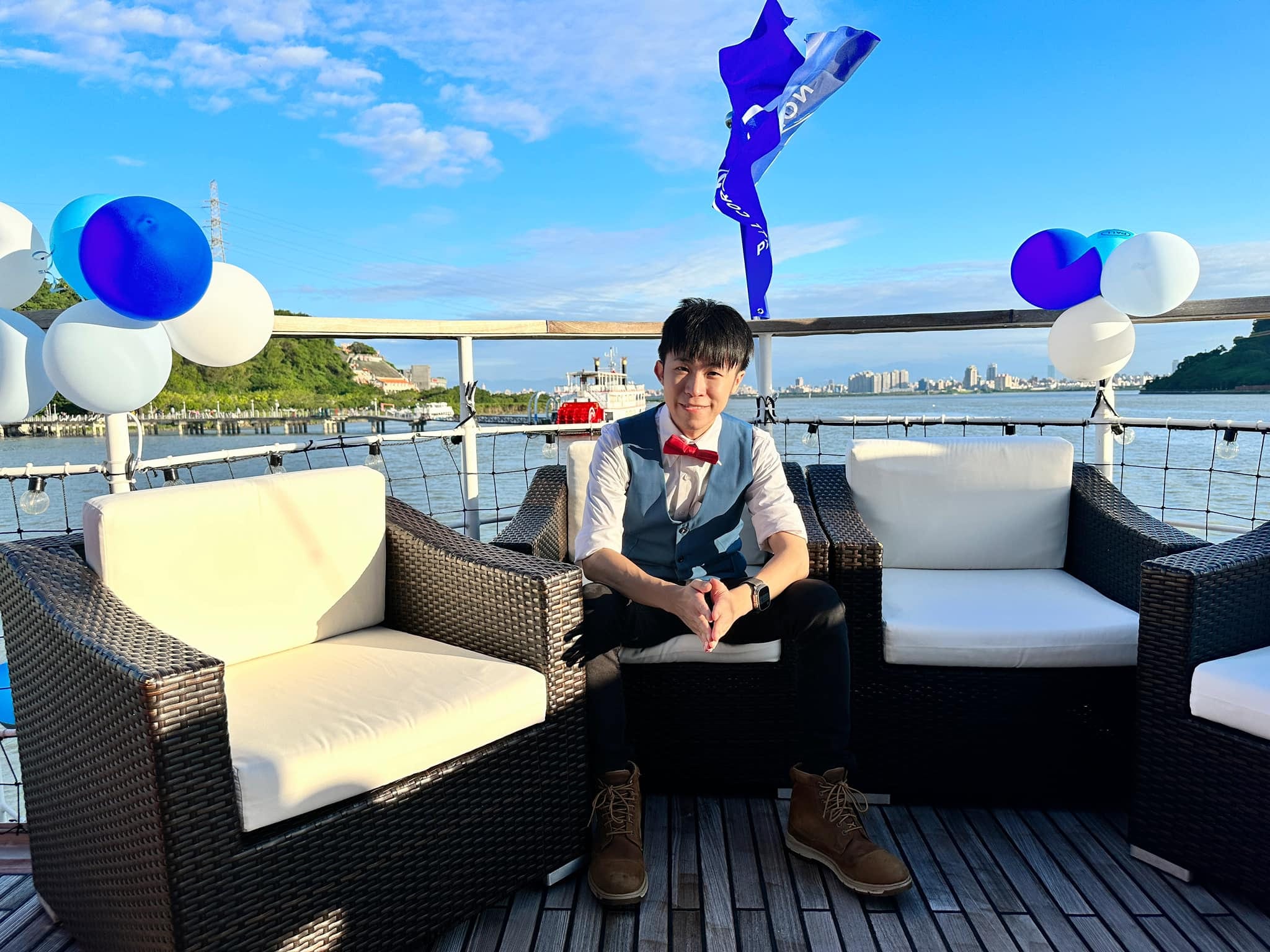 白天的遊艇甲板，柯男坐在白色藤椅上，欄杆綁著藍白色氣球