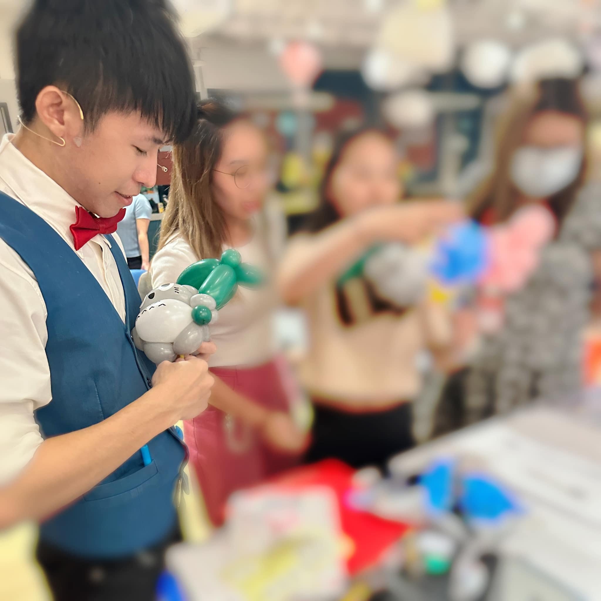 柯男低頭折灰色龍貓頭頂綠葉的造型氣球，身後大家拿著氣球排隊