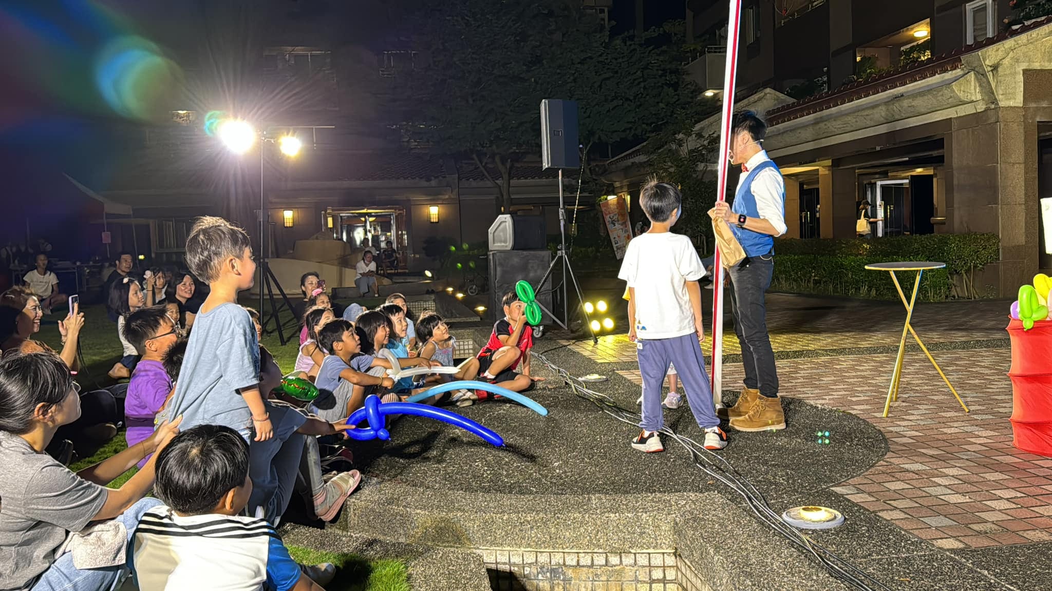 柯男拿著紅白長魔術棒,男孩站在旁邊抬頭看,孩子們圍坐在前方