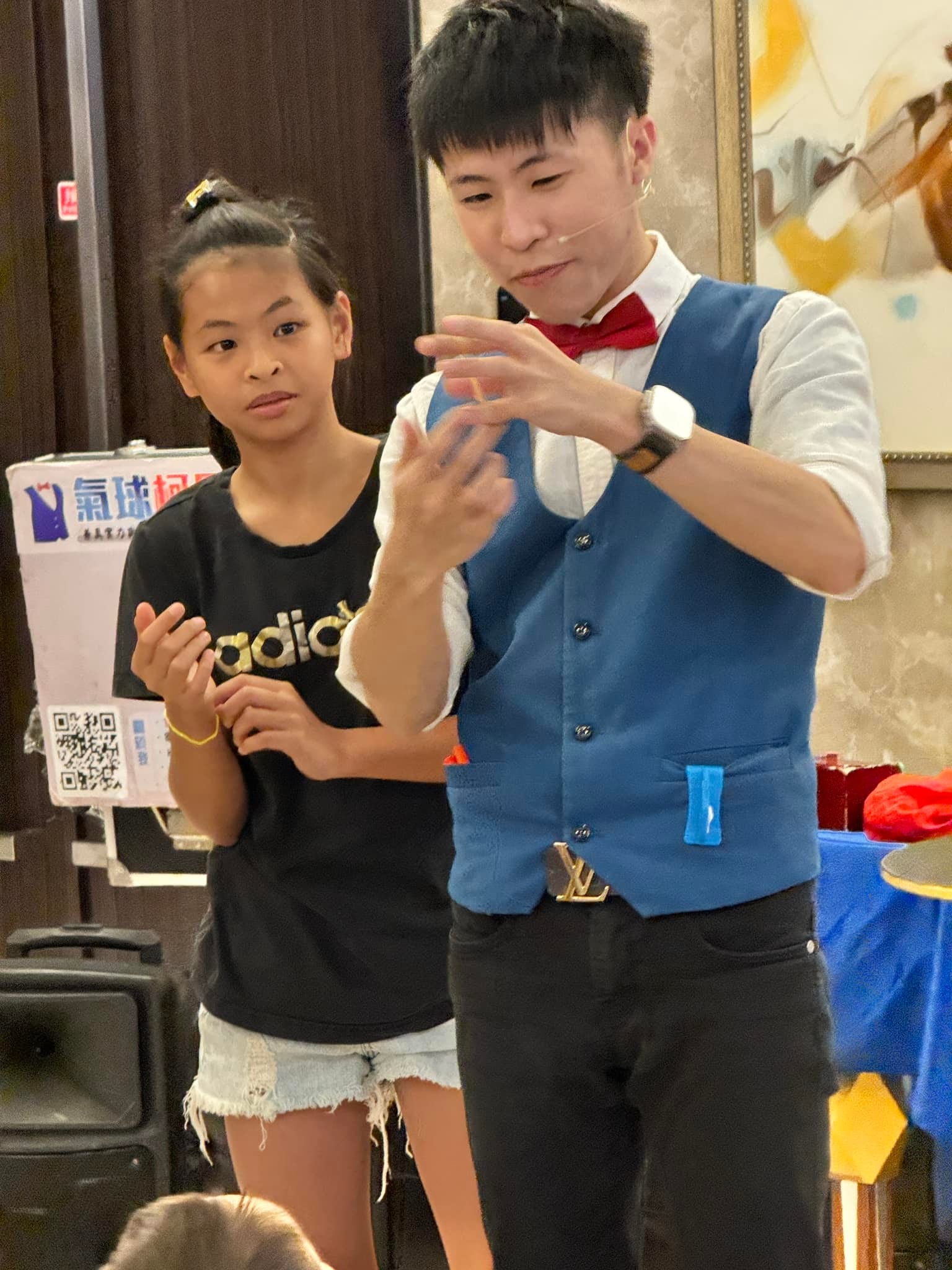 柯男雙手比出魔術手勢,穿黑色上衣的女孩站在旁邊專心看