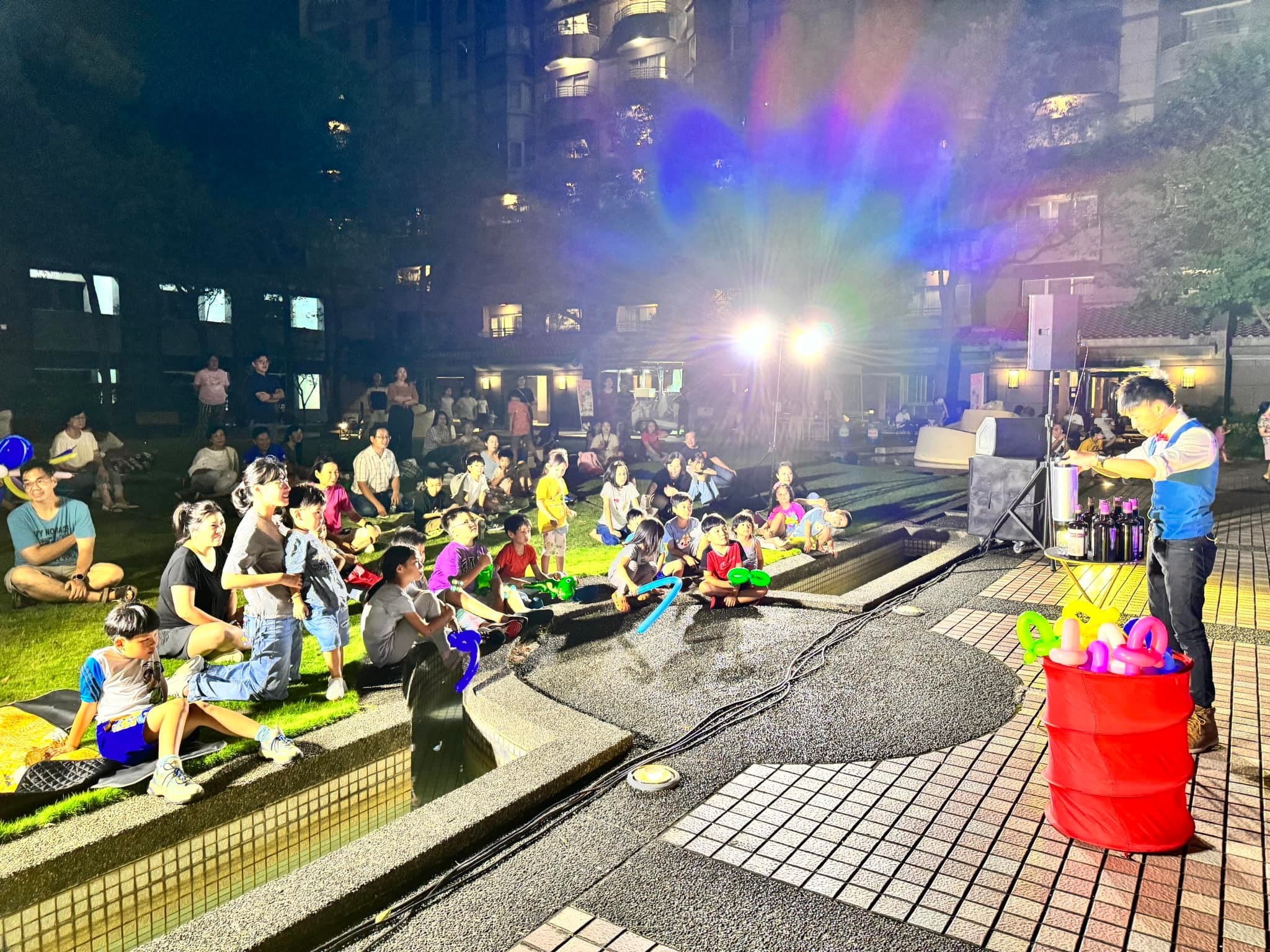 夜晚的社區中庭親子魔術表演,孩子們坐在草地與台階上,柯男站在舞台燈旁