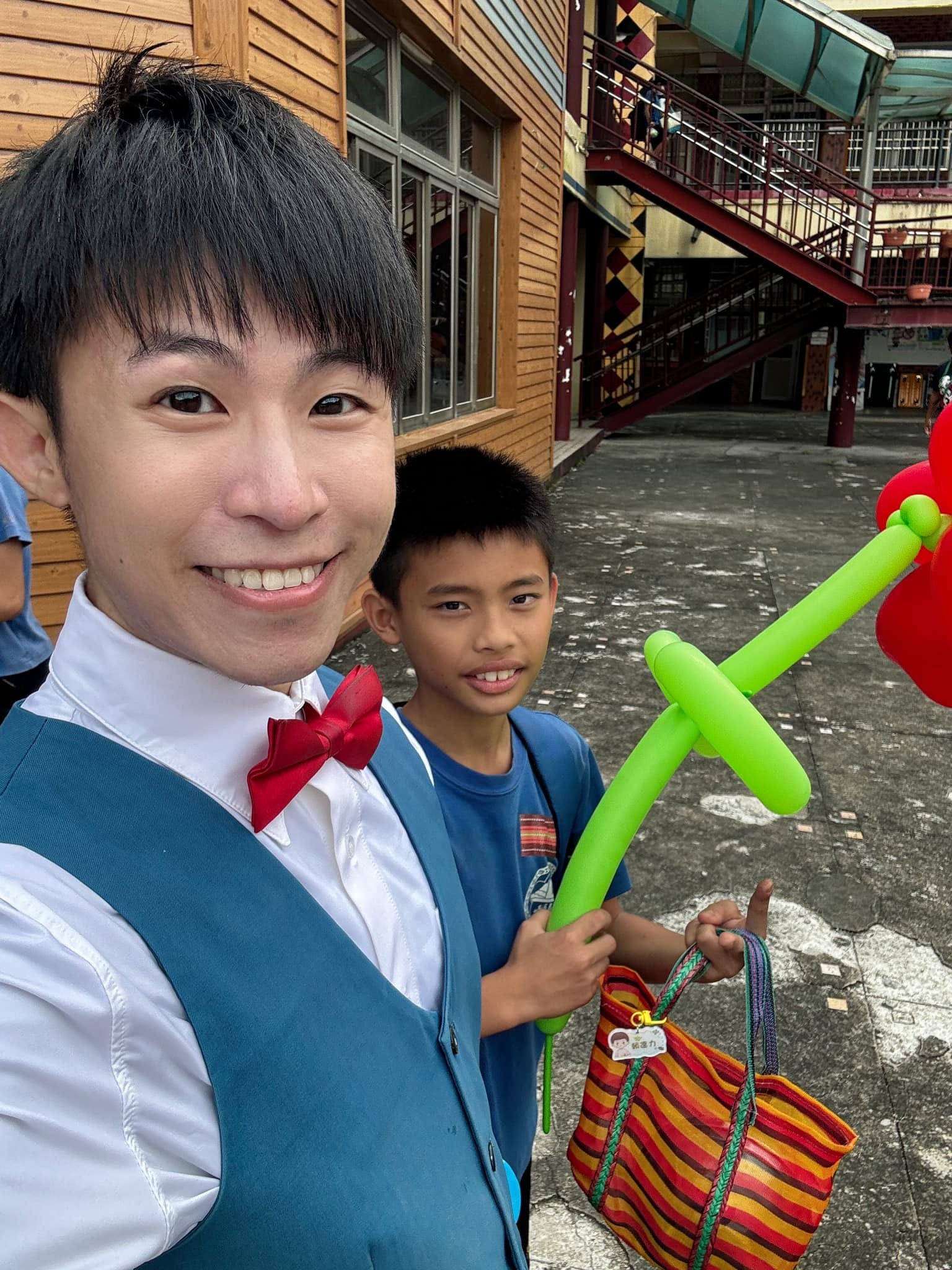 柯男與手拿綠桿紅花氣球的男孩在校舍前自拍，男孩提著彩色條紋袋
