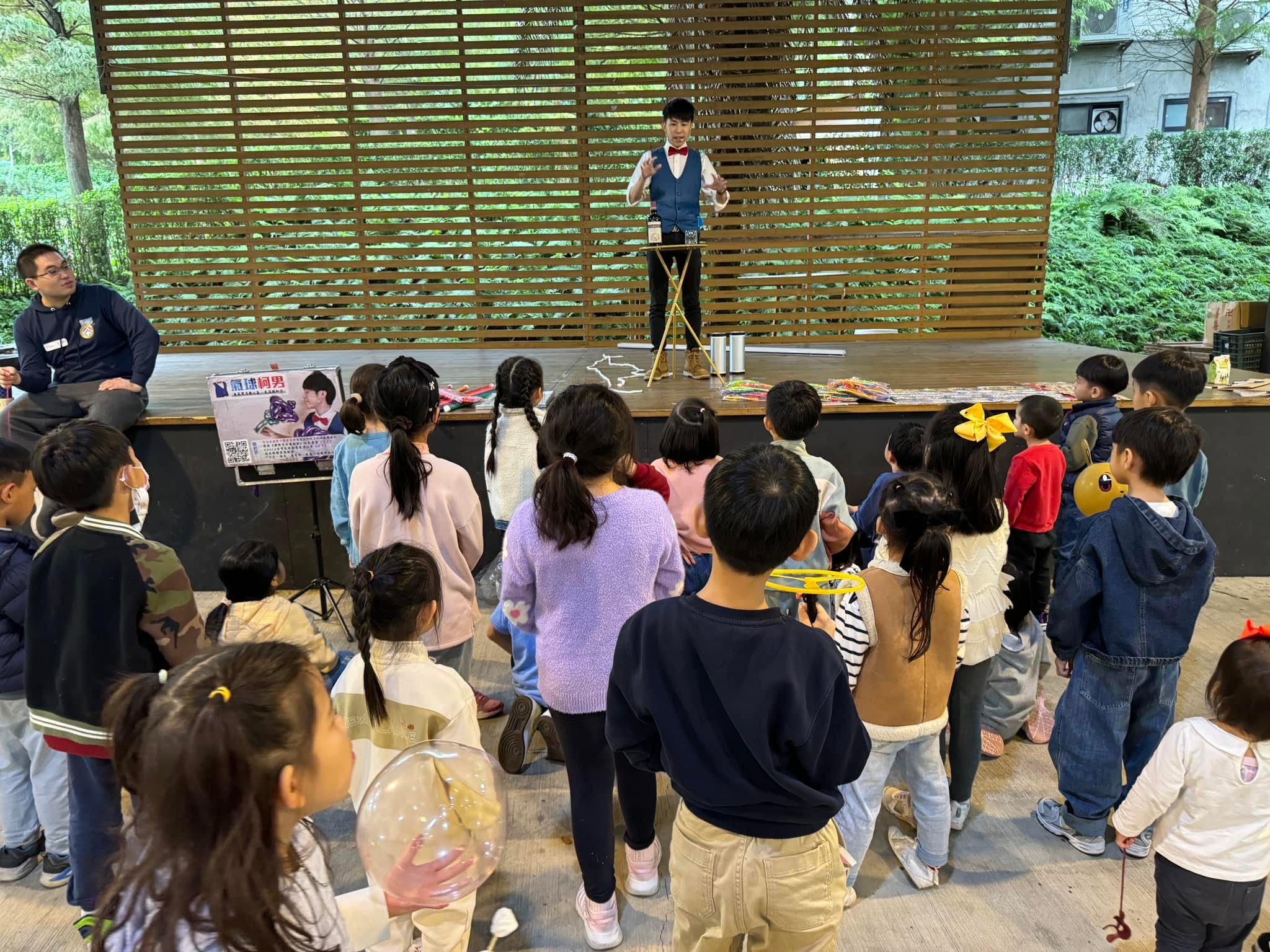 柯男站在木格柵背景的舞台上表演，孩子們站在台前看