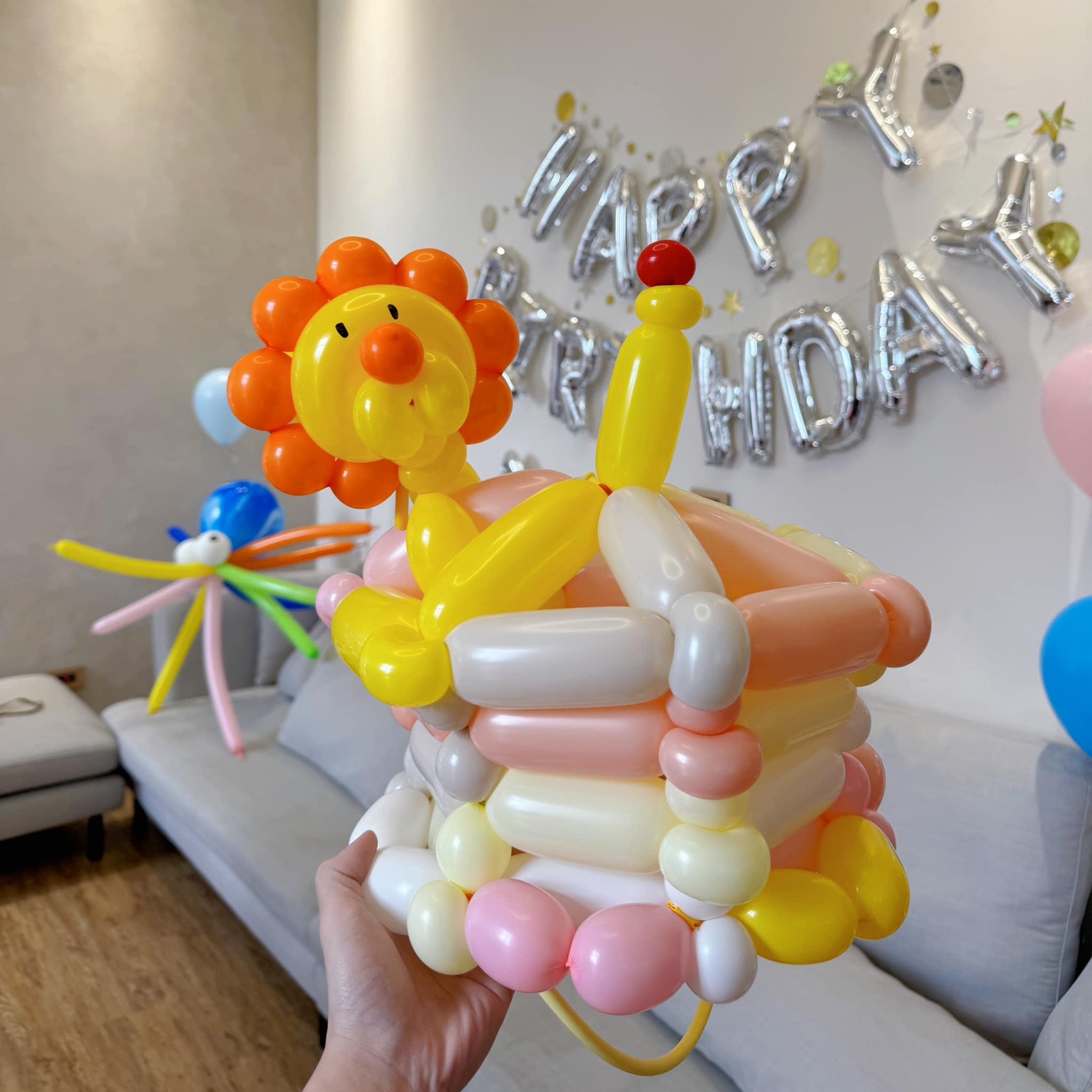 小獅子坐在多層蛋糕上的造型氣球帽，背後是 HAPPY BIRTHDAY 字母