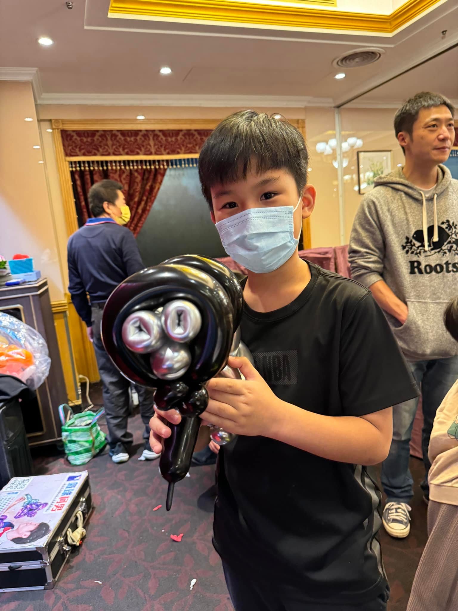 宴會廳裡戴口罩的小男生拿著黑色握把、黑框包著三個銀色圓筒的鍍鉻造型氣球，後方是紅色布幔