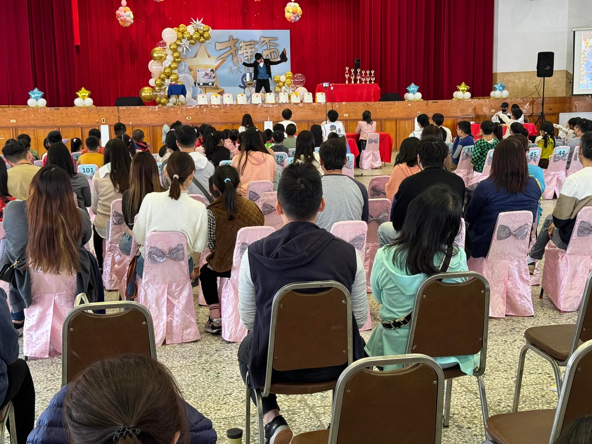 禮堂裡家長和孩子坐在粉紅椅套座位上，看台上魔術表演