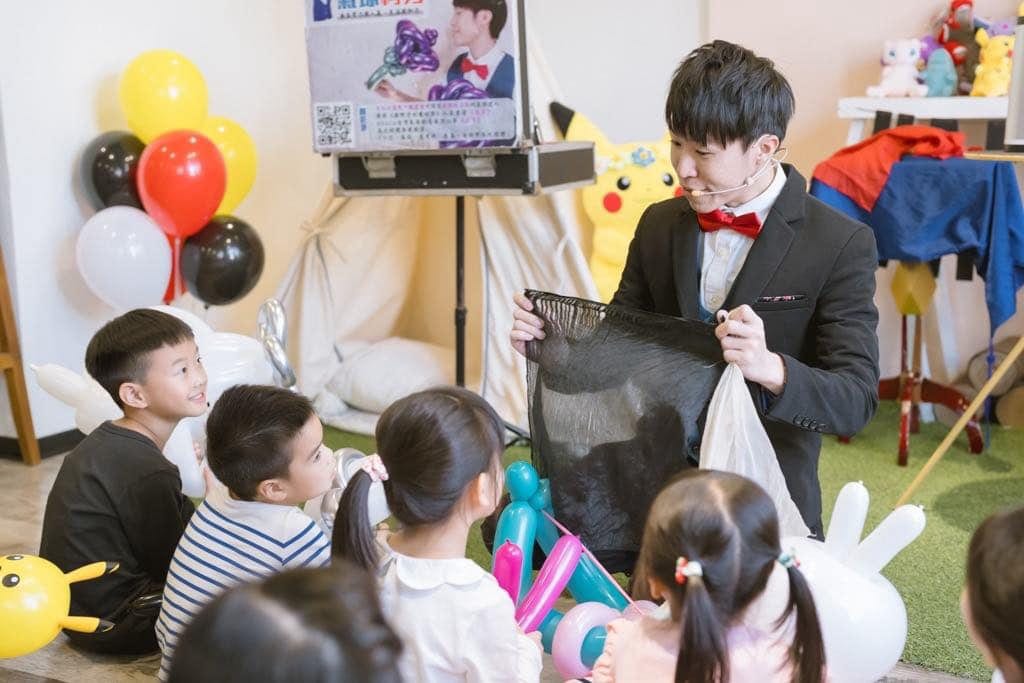 柯男拿著黑色魔術袋表演，孩子們坐在前方仰頭看