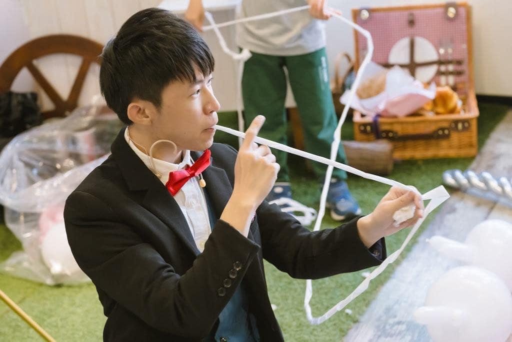 柯男咬著白色繩子表演繩子魔術，背景是野餐風道具