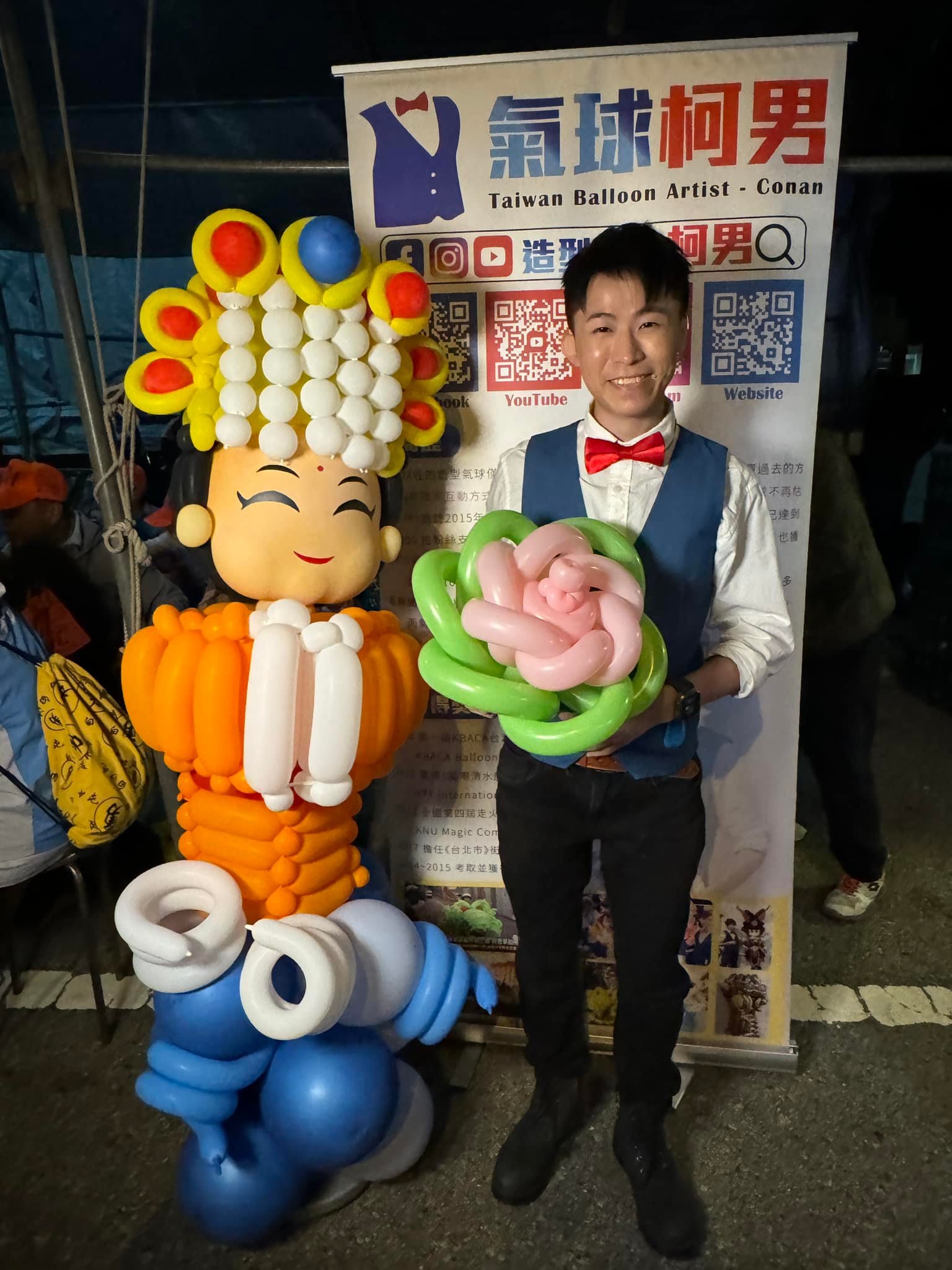 柯男拿著粉紅綠色花朵氣球，站在媽祖氣球與看板之間