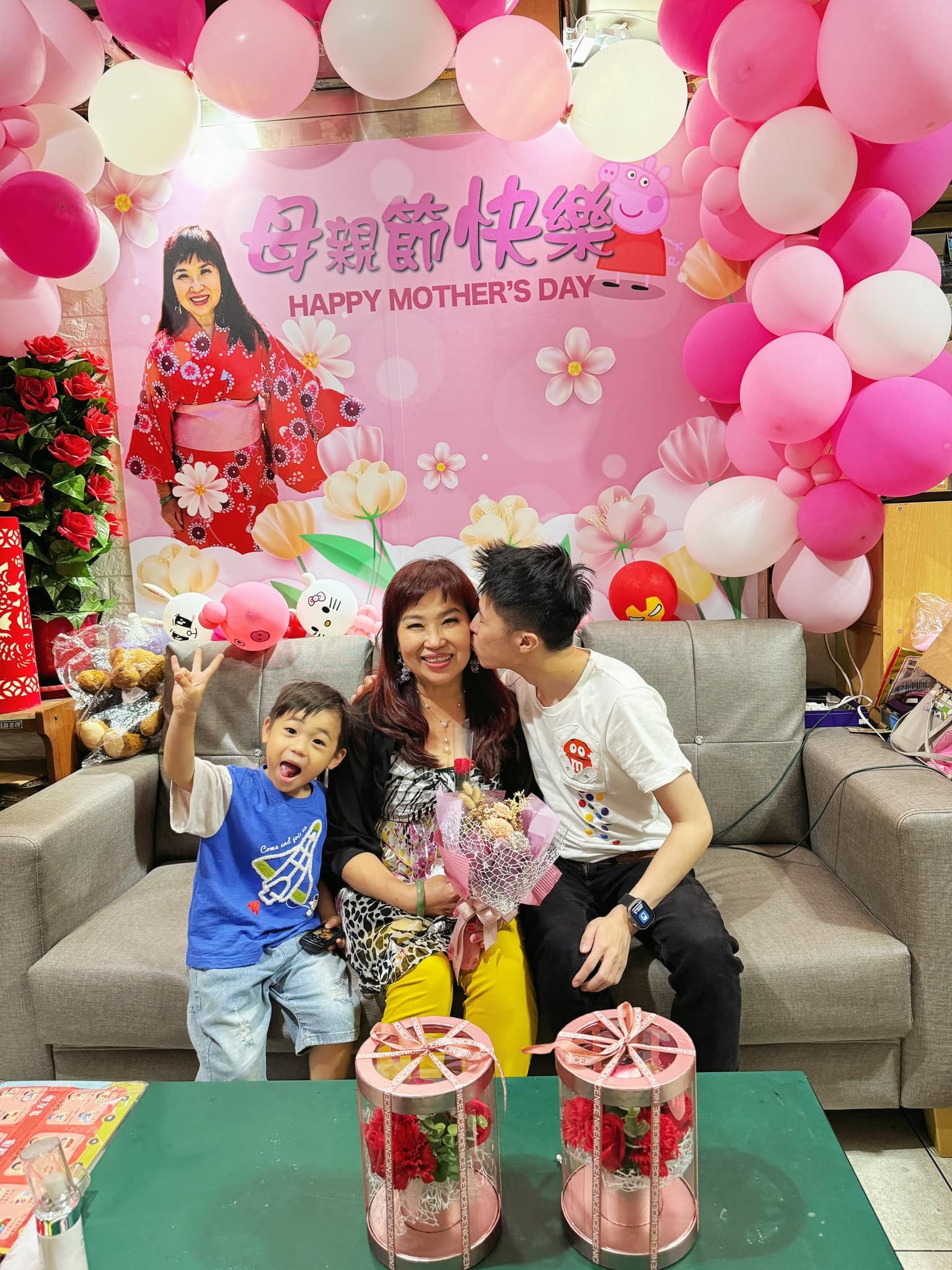 印著母親節快樂、媽媽照片與佩佩豬的粉紅背板，兩側桃紅粉白氣球拱，我親媽媽臉頰，外甥坐在旁邊比 YA