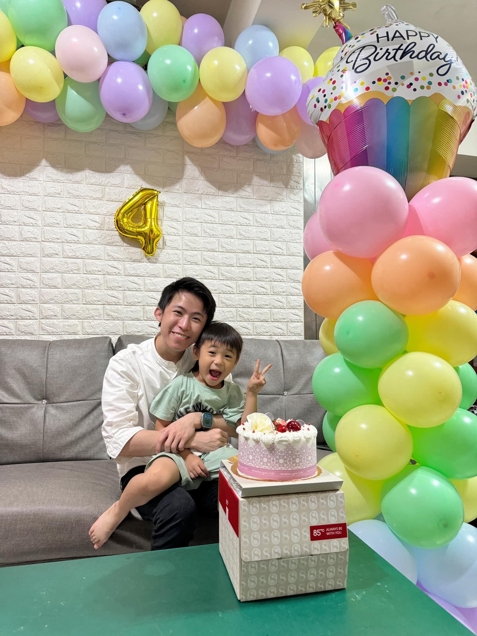白磚牆上的馬卡龍色氣球串與金色數字 4 氣球，右側彩色氣球柱頂著生日杯子蛋糕鋁箔氣球，我抱著外甥坐在沙發上