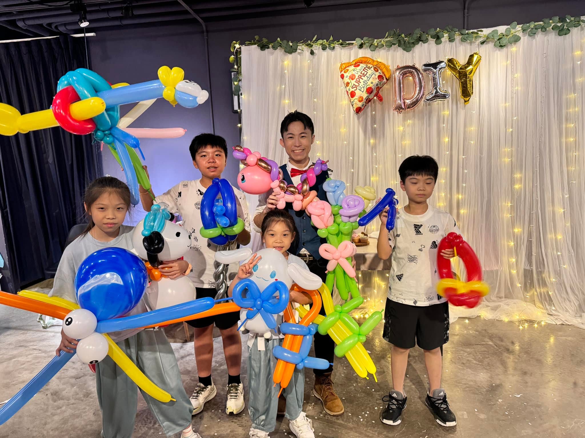 孩子們拿著寶劍、大耳狗、花束等氣球，站在掛著 DIY 字母氣球的白紗背板前