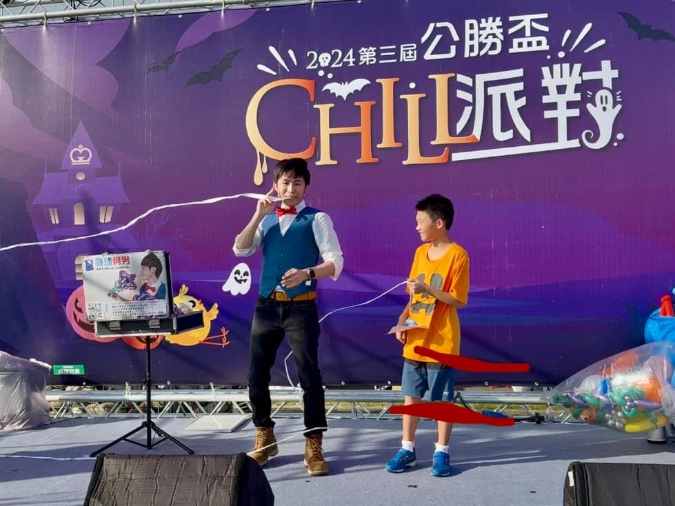 戶外舞台上，柯男和一位穿橘色上衣的男孩拉著白色繩子表演魔術