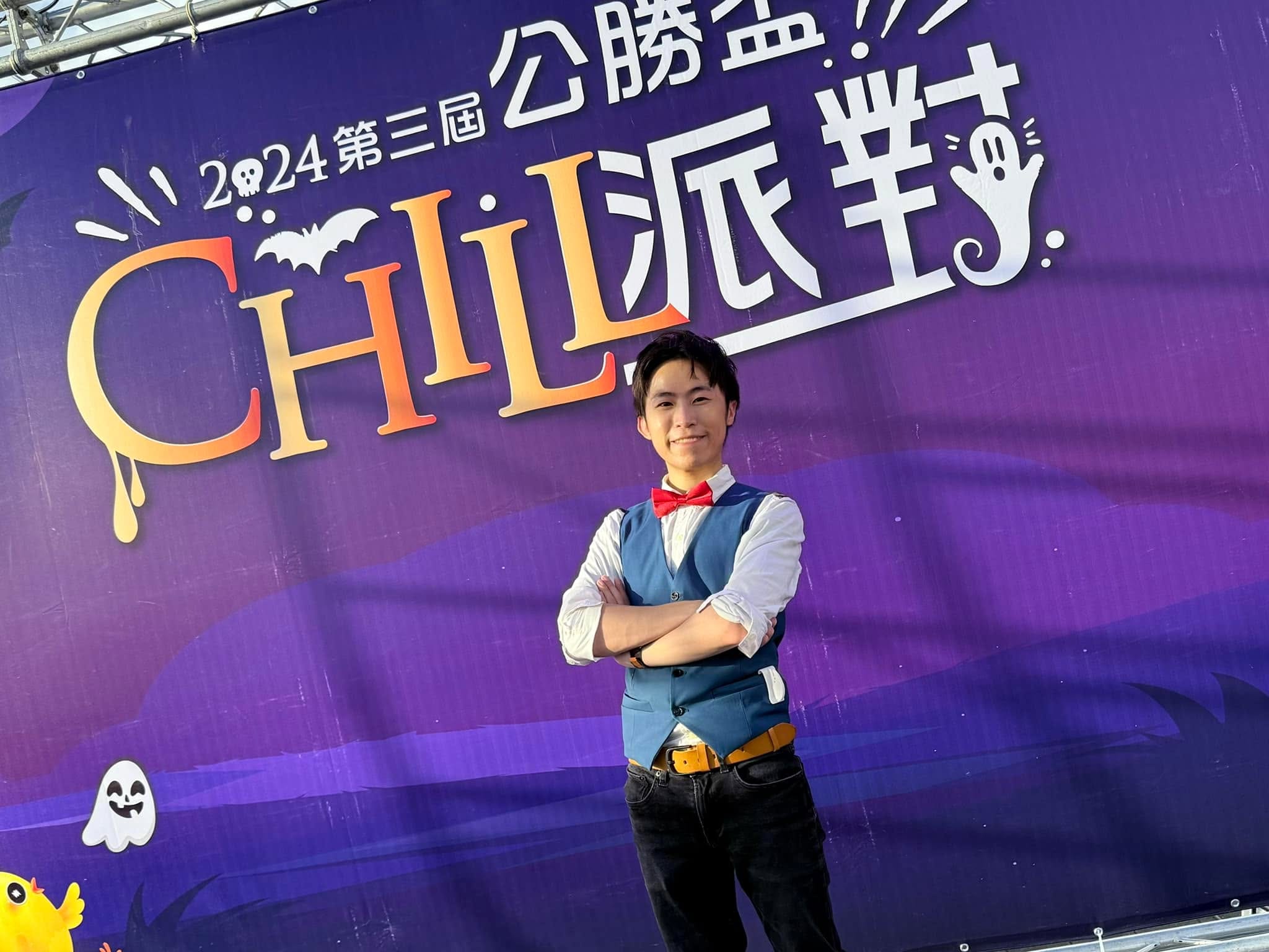 柯男雙手抱胸站在萬聖節派對紫色主視覺背板前，背板上有蝙蝠與幽靈圖案