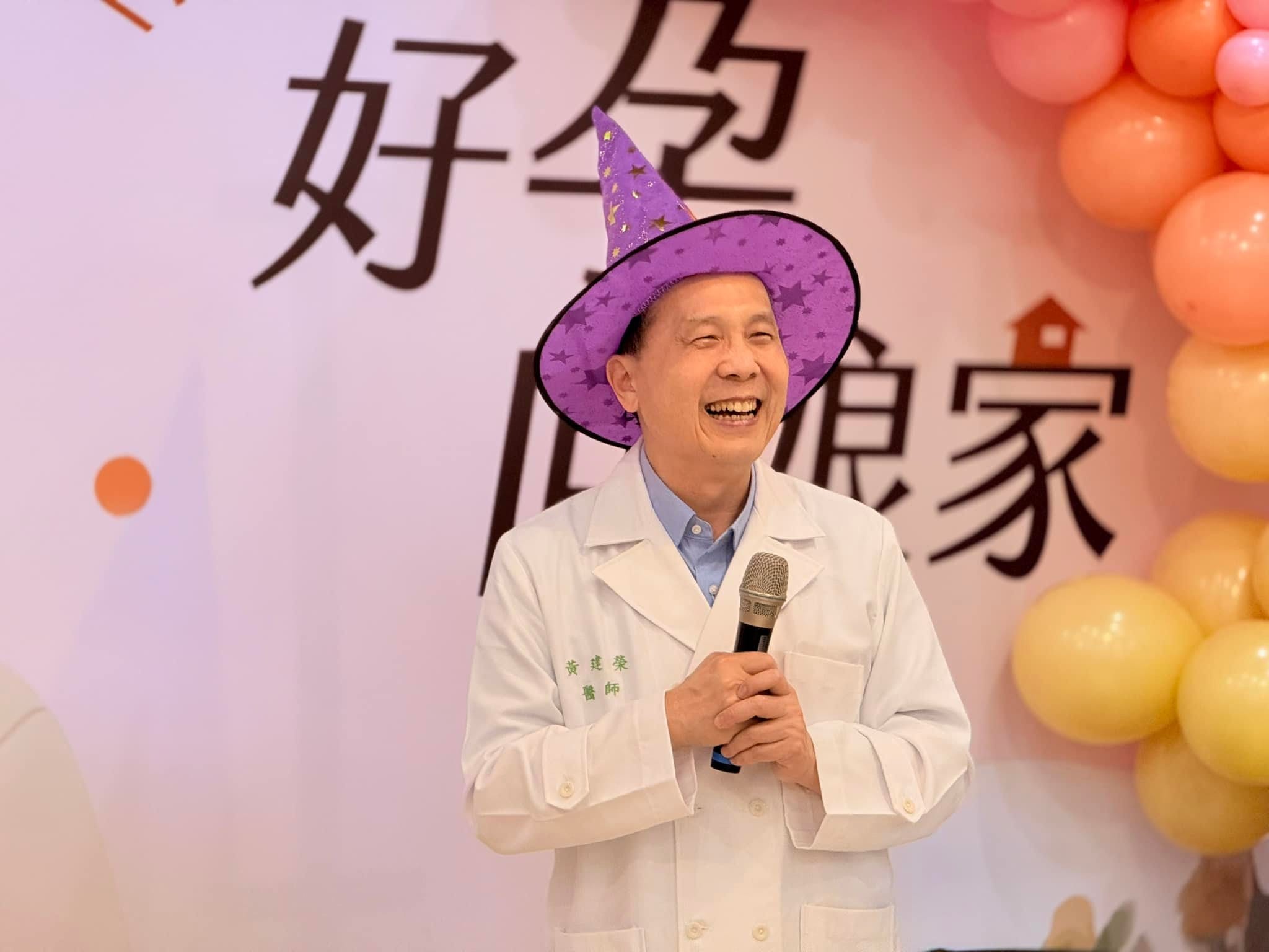 戴紫色巫師帽、穿白袍的院長拿著麥克風在氣球花圈前大笑