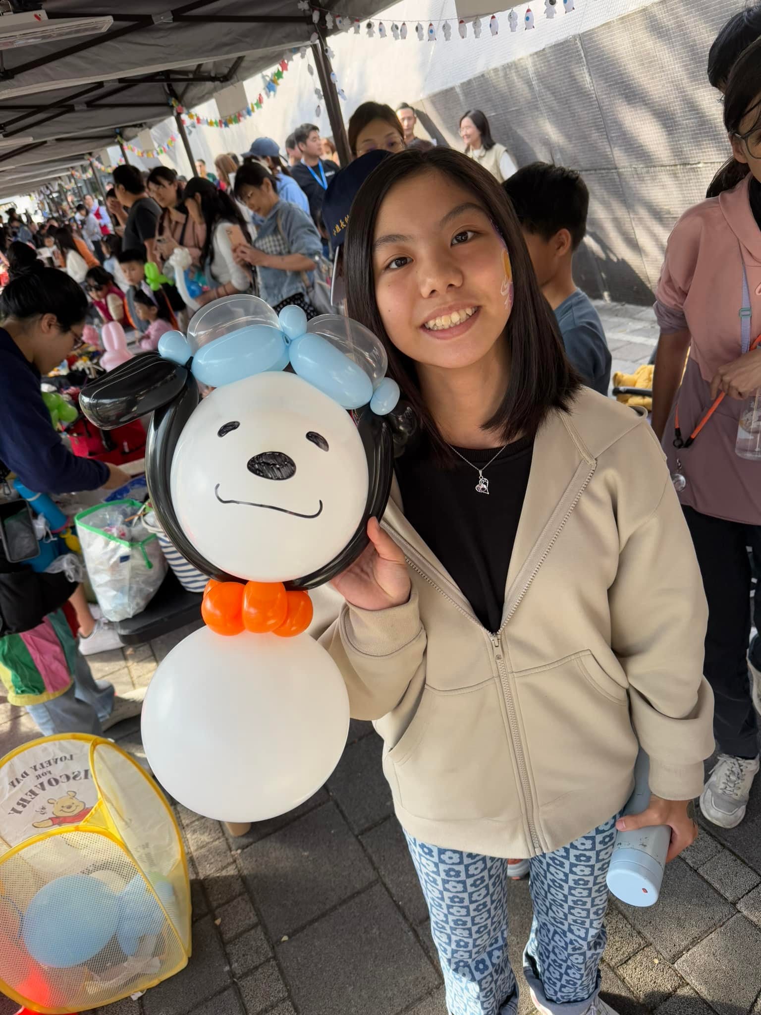 女孩捧著戴藍色飛行帽、橘色圍巾的史努比造型氣球，背景是藝術節市集帳篷