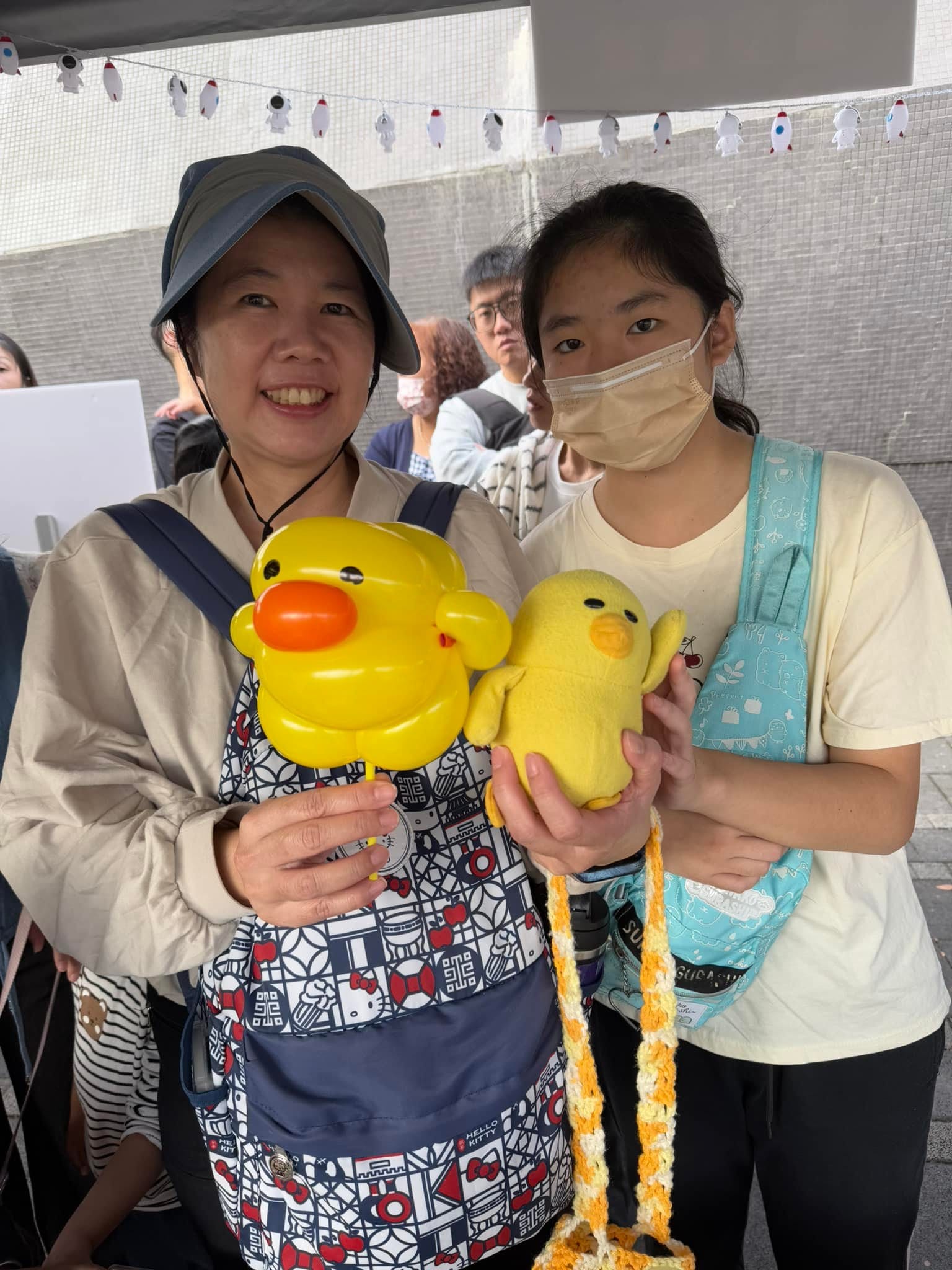 媽媽拿著黃色小鴨造型氣球，旁邊的女兒拿著同款黃色小鴨玩偶