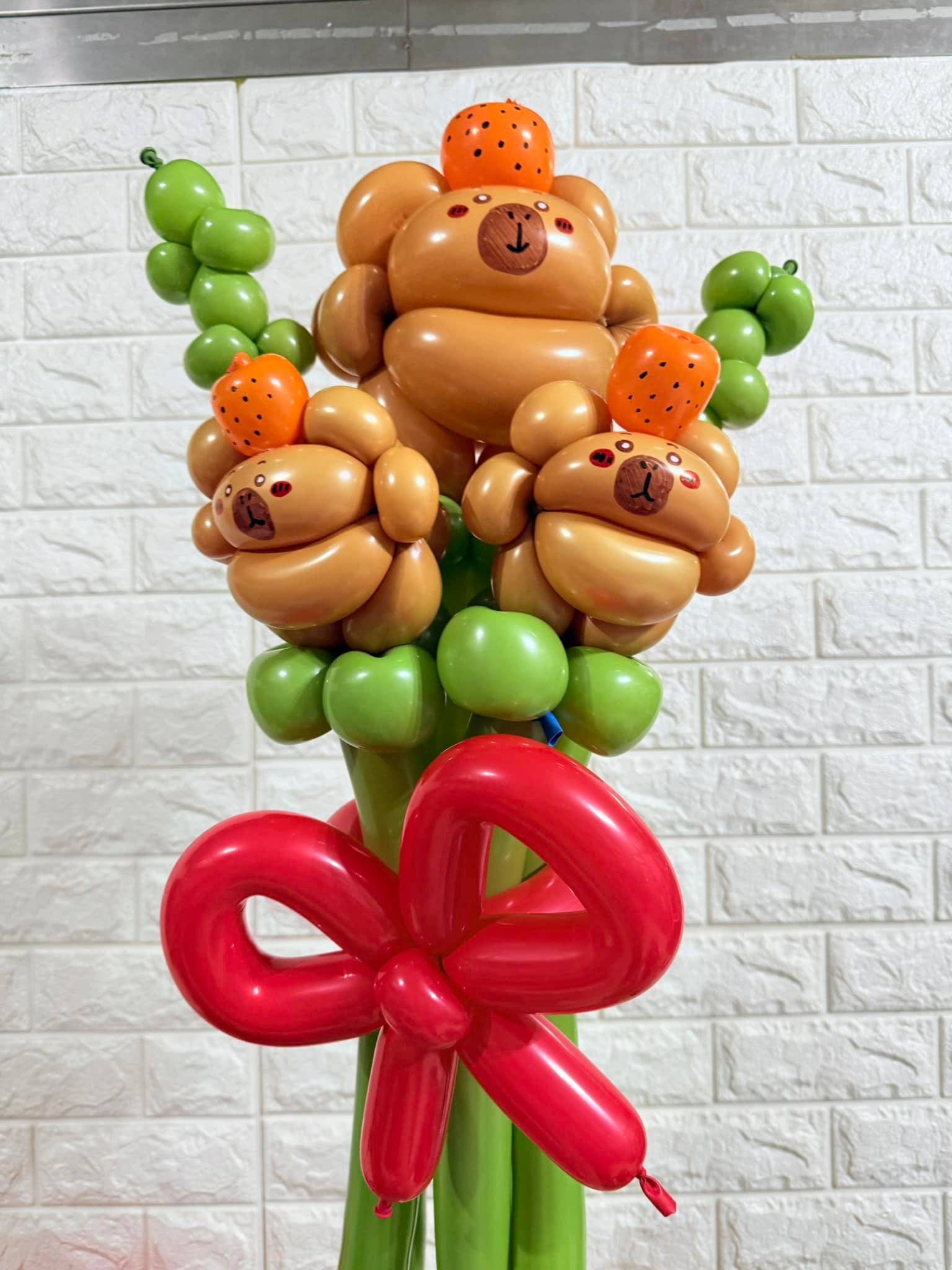 三隻頭頂橘子的卡比巴拉造型氣球組成的花束，搭配綠色葉子與紅色蝴蝶結