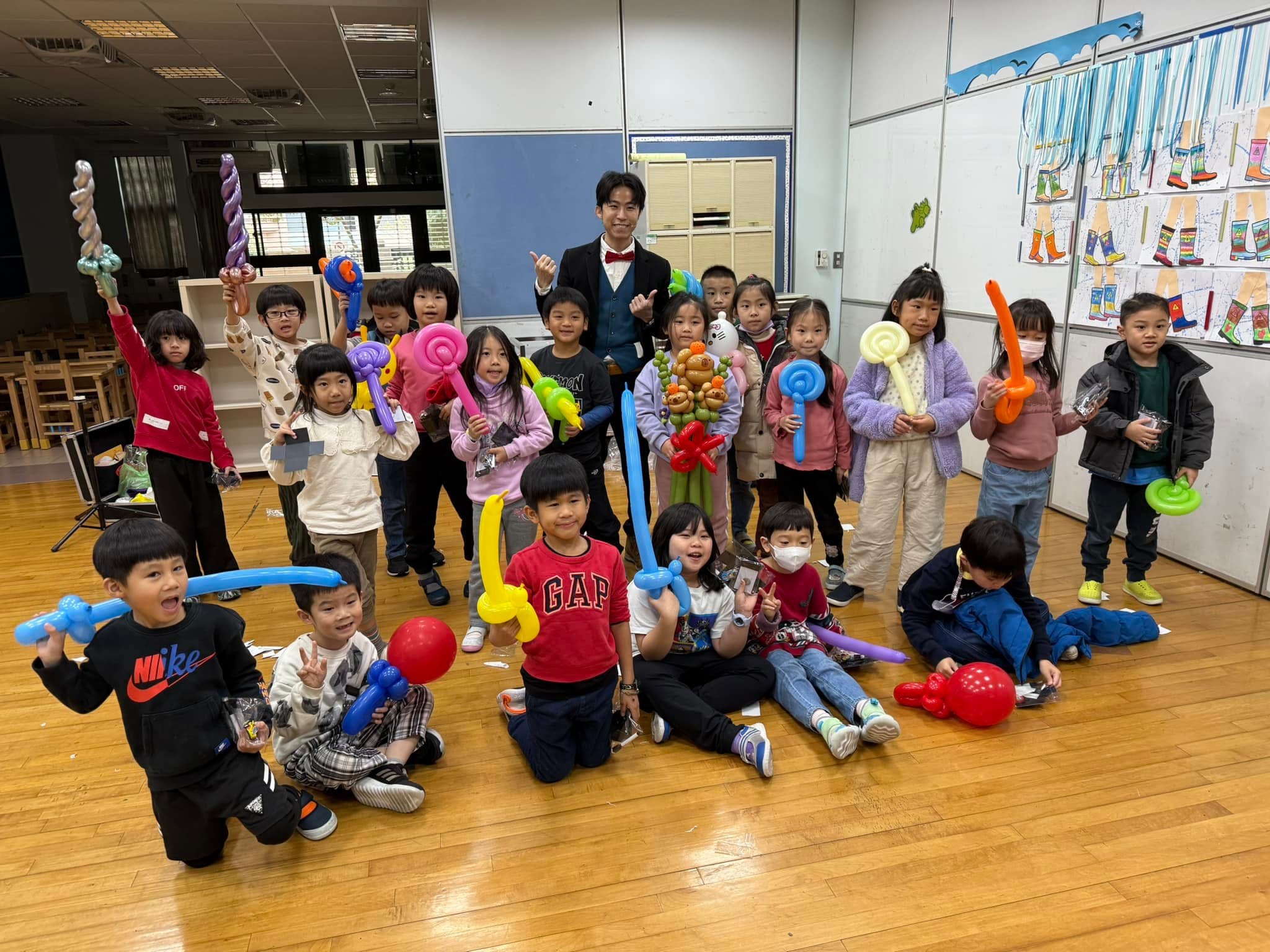 教室木地板上，全班小朋友拿著寶劍、小狗等造型氣球和柯男合照