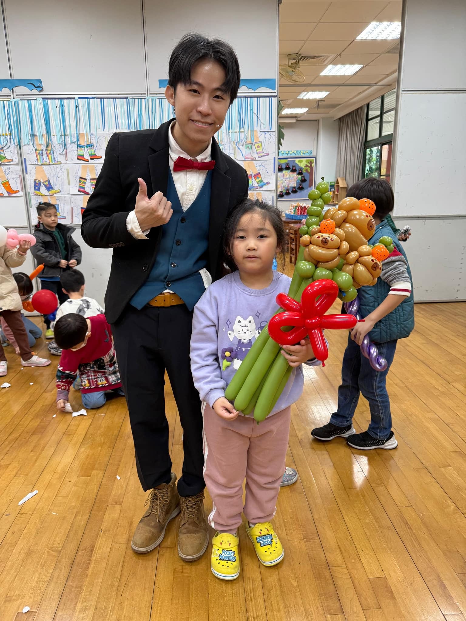 穿淺紫色上衣的小女孩在教室裡抱著卡比巴拉氣球花束，柯男在旁比讚