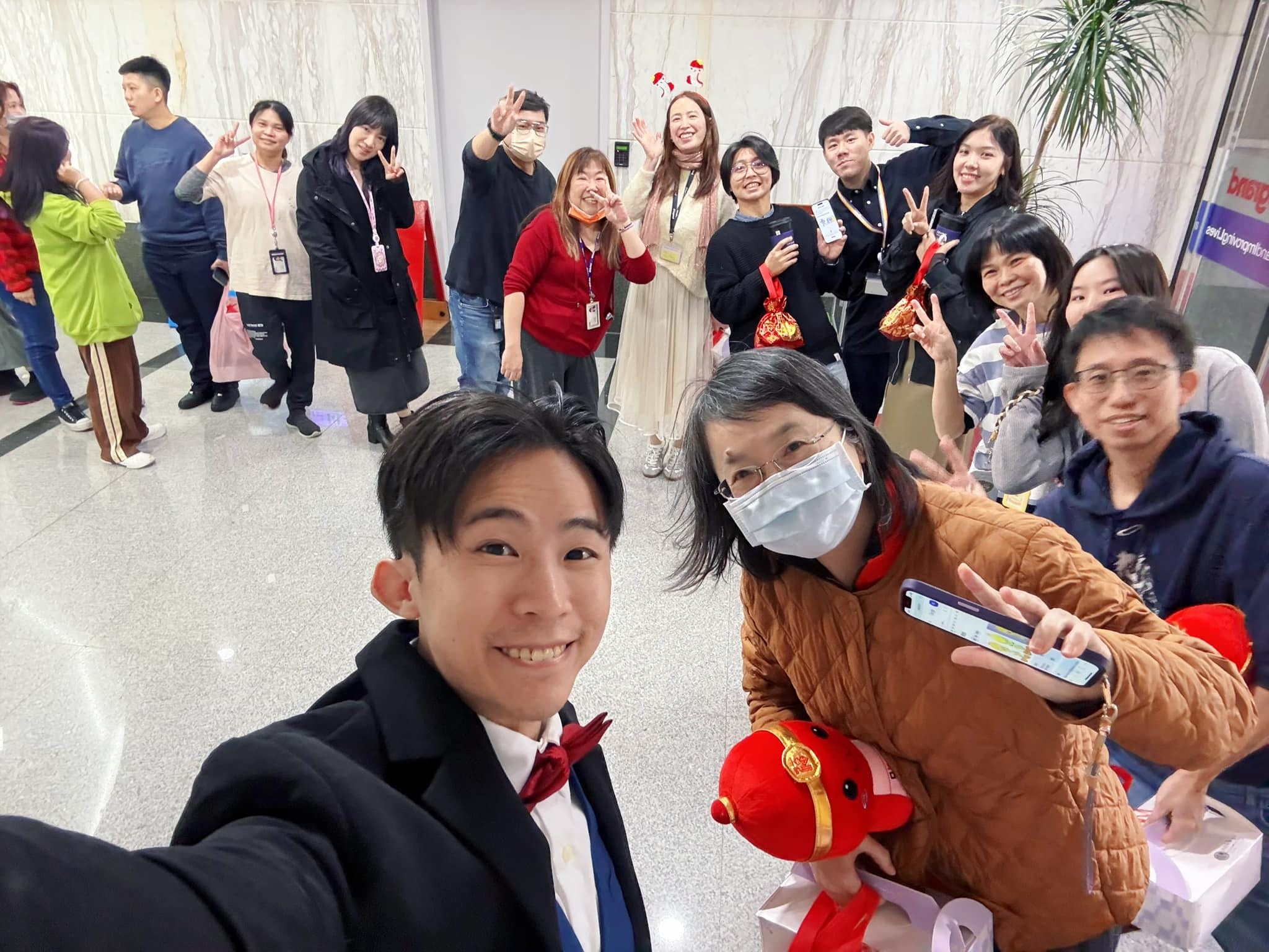 柯男在辦公大樓大廳和一群拿著紅色福袋的同仁自拍，大家比 YA 開心合照