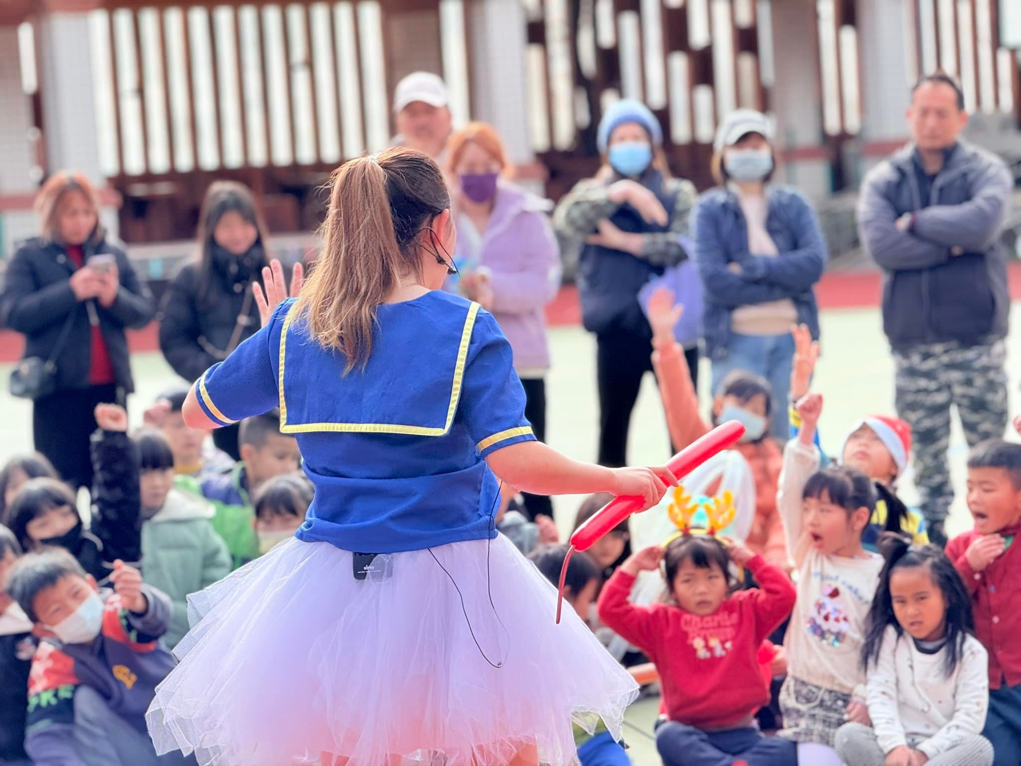 穿藍色水手服、白色紗裙的 Eva 背對鏡頭拿著紅色長條氣球，面前是坐在操場上的孩子與家長