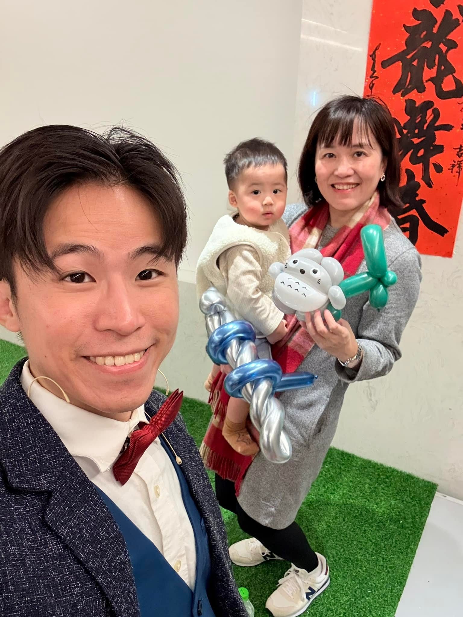 柯男和抱著小男孩的媽媽自拍，小男孩拿著灰白色角色與銀藍色造型氣球
