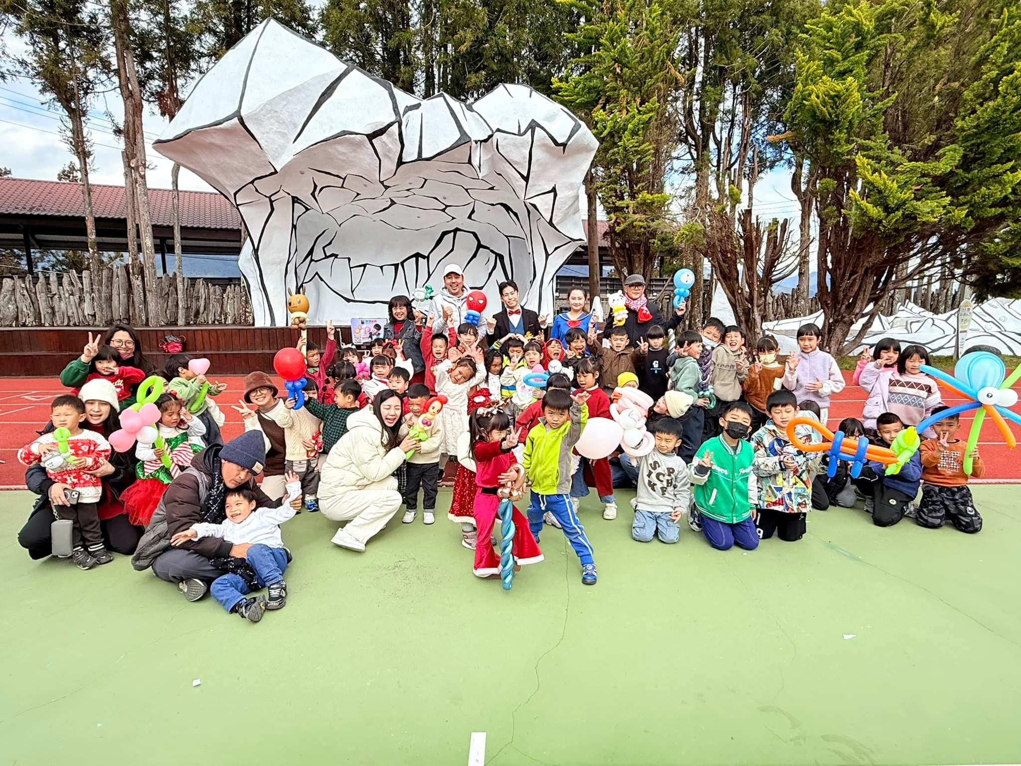 台中梨山小學操場上，師生與家長拿著造型氣球大合照，背景是白色岩石造型牆與高山樹林