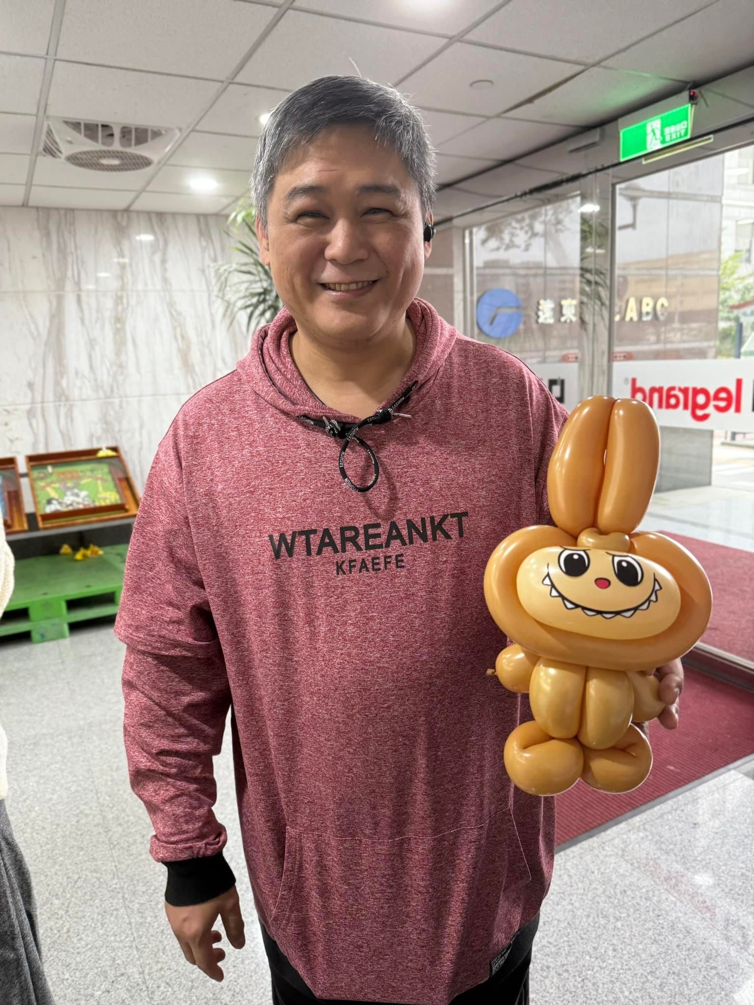 穿紅色連帽衣的男士拿著咖啡色 Labubu 造型氣球，在大廳門口微笑