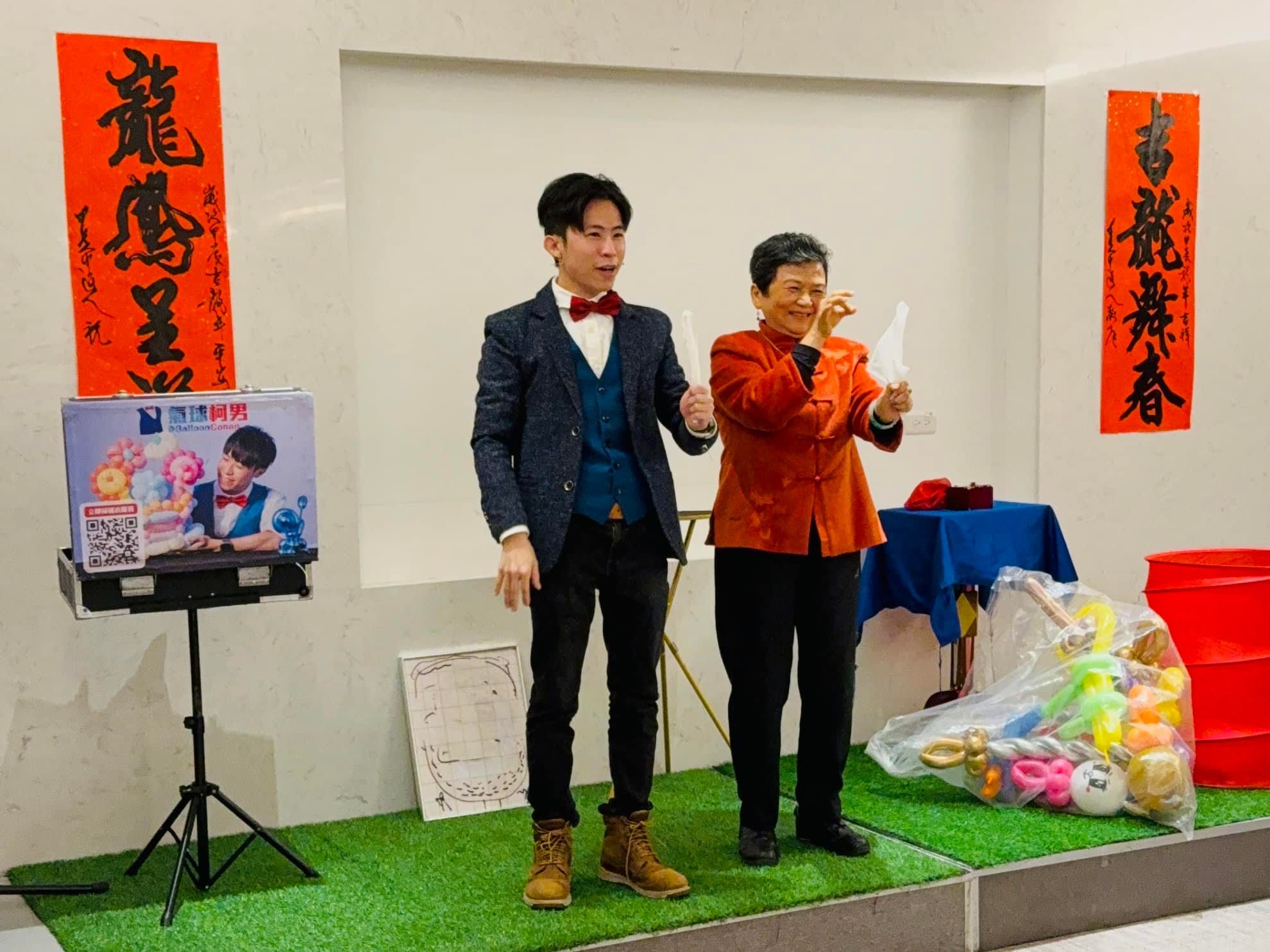 柯男在交誼廳舞台上和一位穿橘紅外套的長輩一起拿白色道具表演魔術，牆上貼著春聯