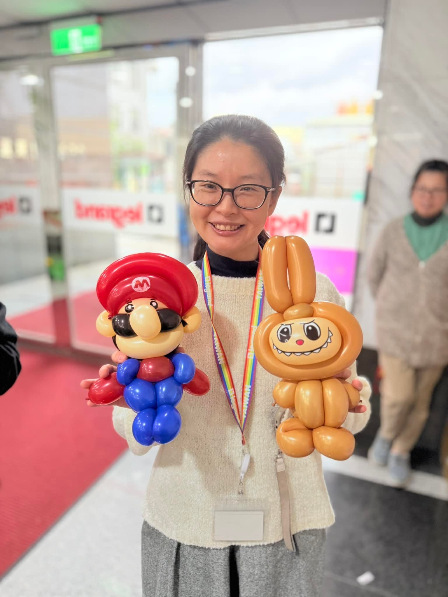 戴眼鏡的女士左手拿瑪利歐、右手拿 Labubu 造型氣球，開心看鏡頭