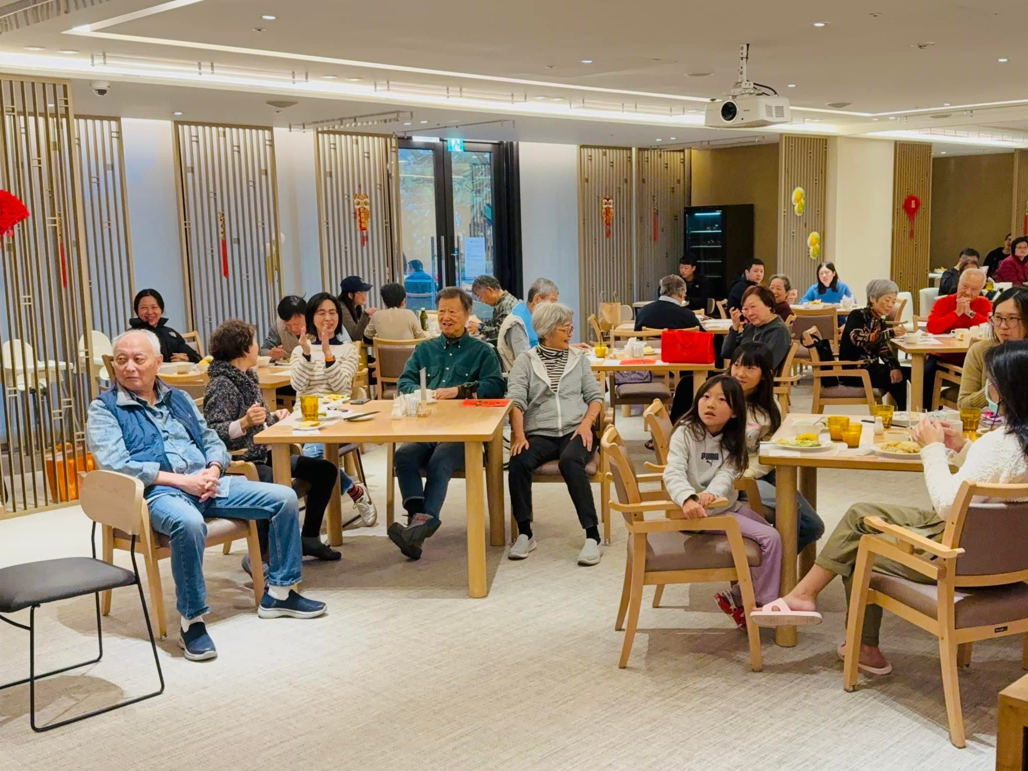 酒店式公寓交誼廳裡，長輩與家人坐在木桌旁觀看表演，天花板掛著紅色燈籠