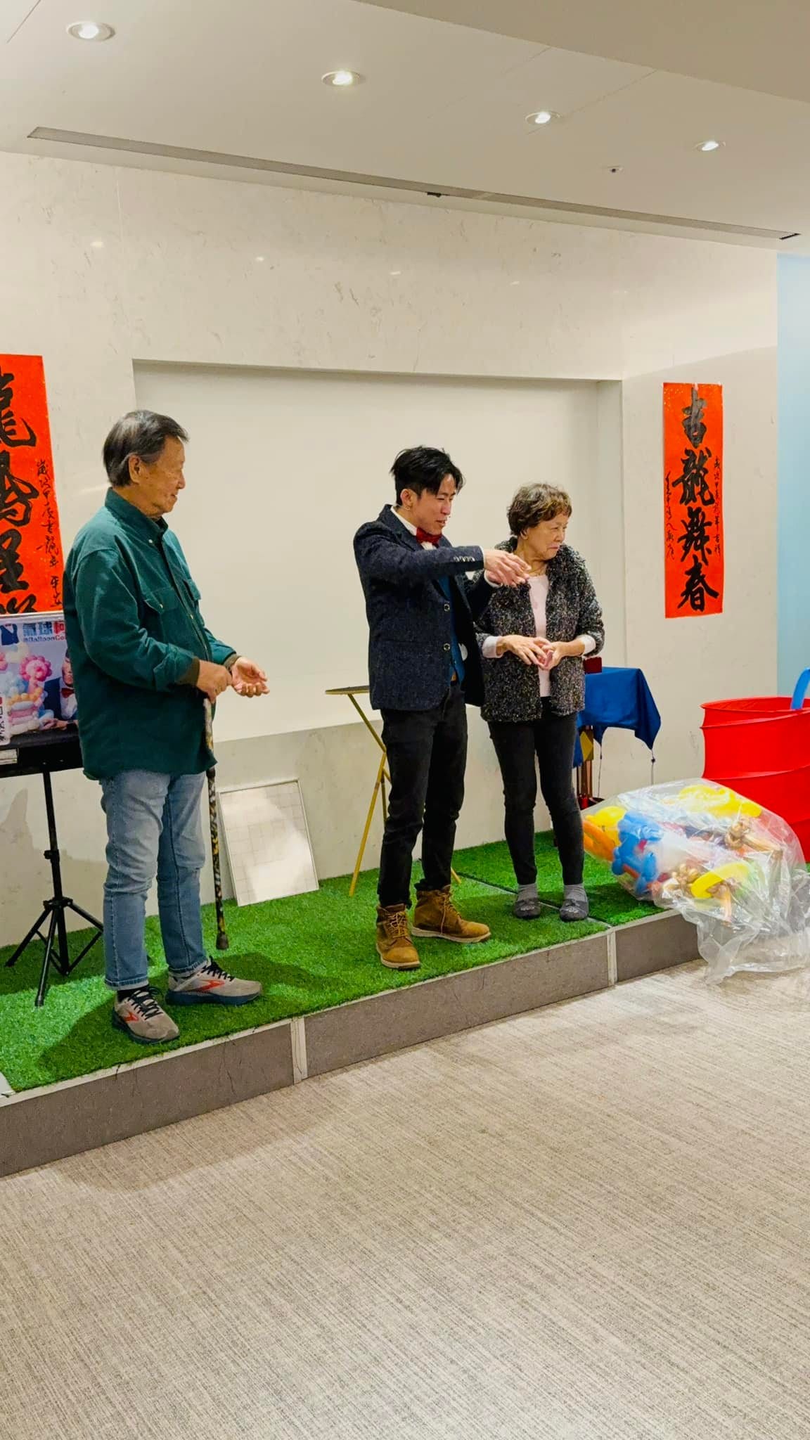 人工草皮舞台上，柯男請一位阿姨上台協助，旁邊拄拐杖的北北一起看魔術