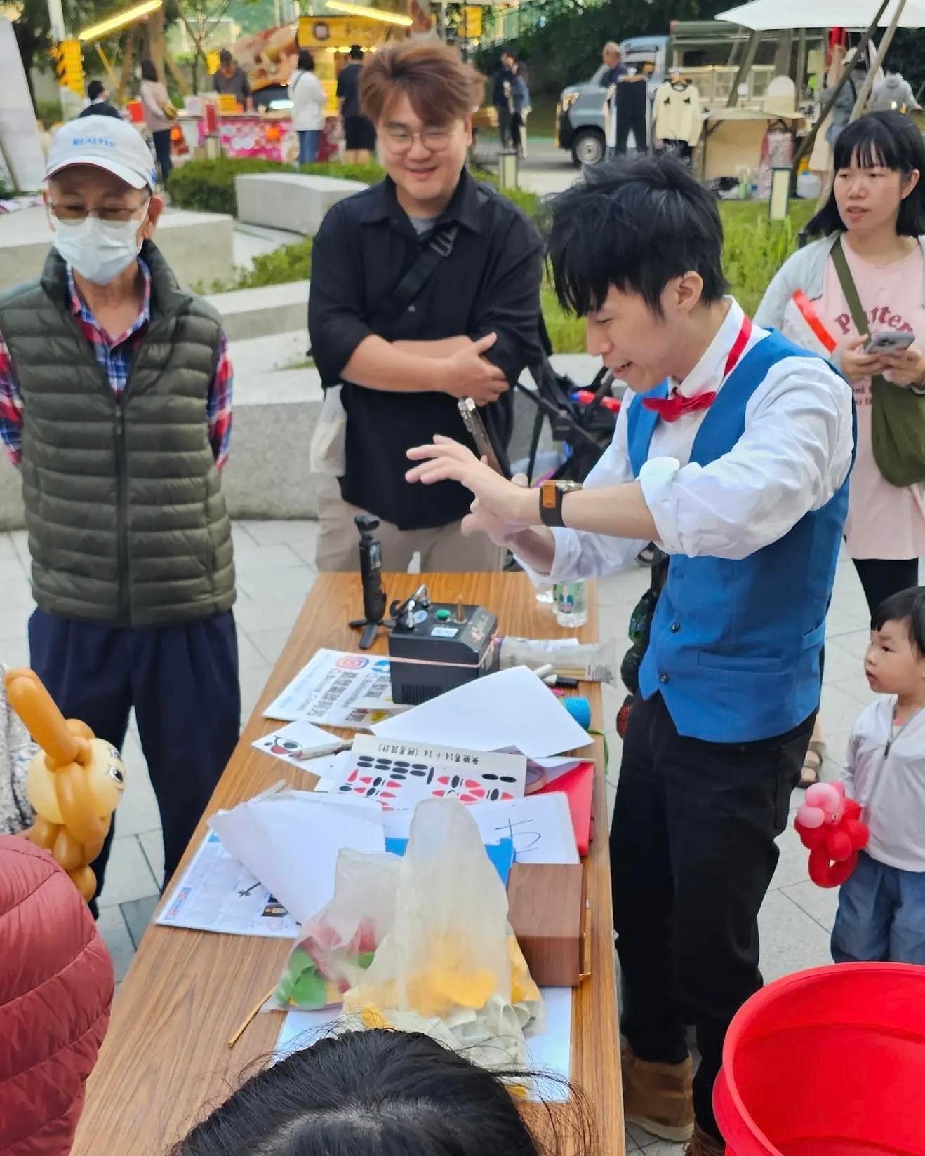 社區市集廣場上，柯男在木桌前現場折氣球，周圍站著排隊的大人和小朋友