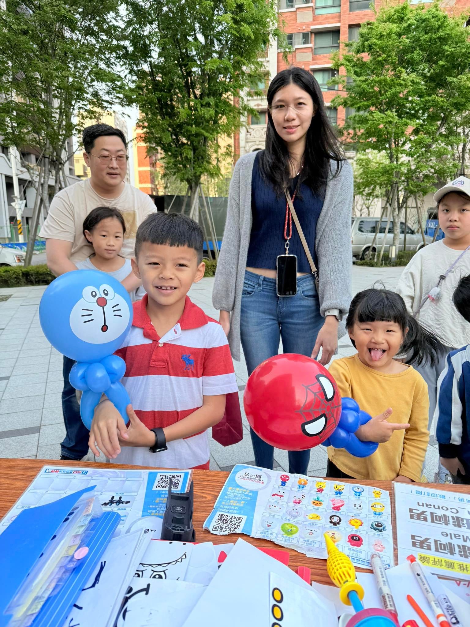 社區市集攤位前，男孩拿著哆啦A夢造型氣球，女孩拿著蜘蛛人造型氣球吐舌頭