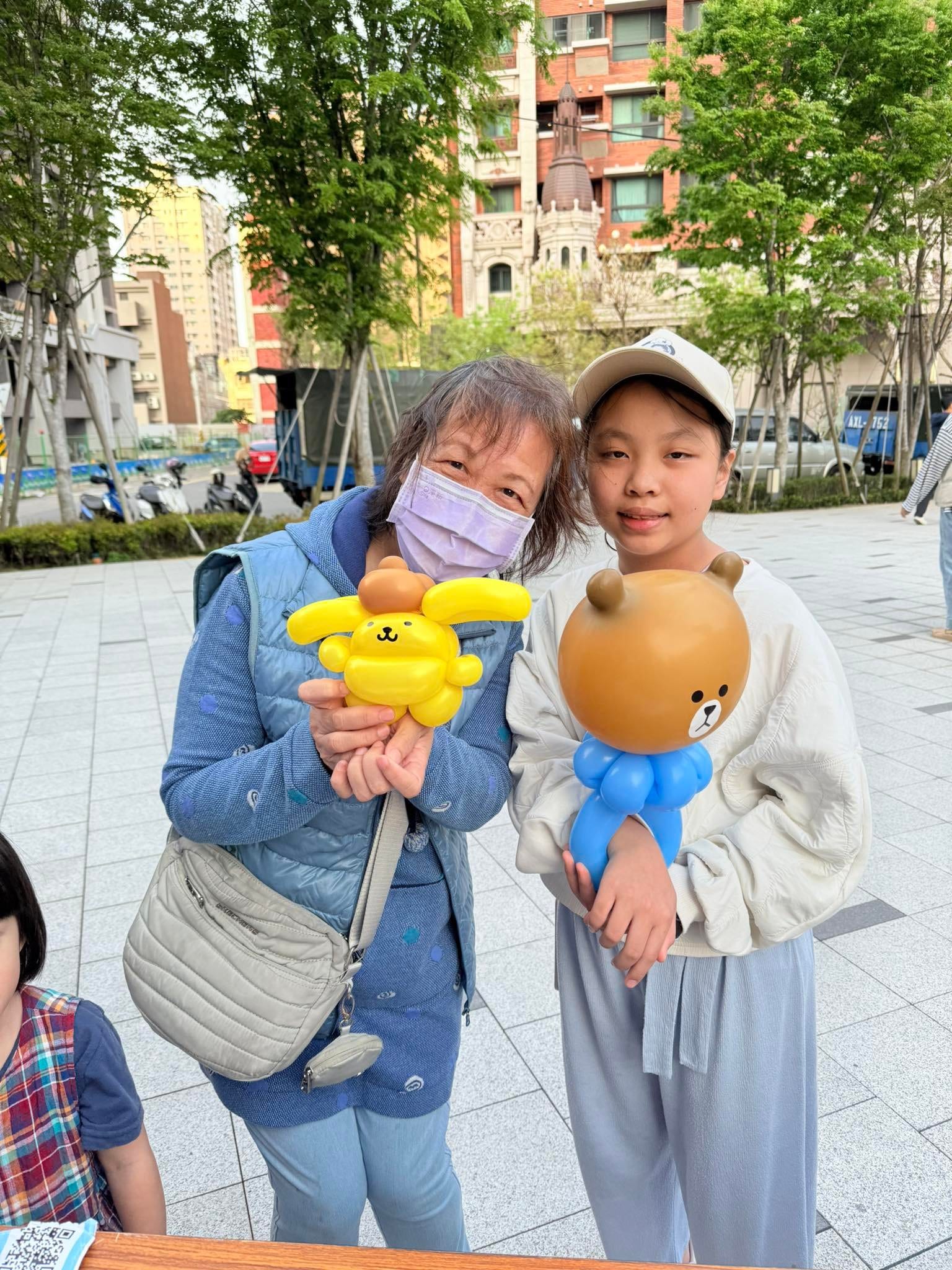 奶奶和女孩站在一起，分別拿著戴咖啡色帽子的布丁狗和熊大造型氣球