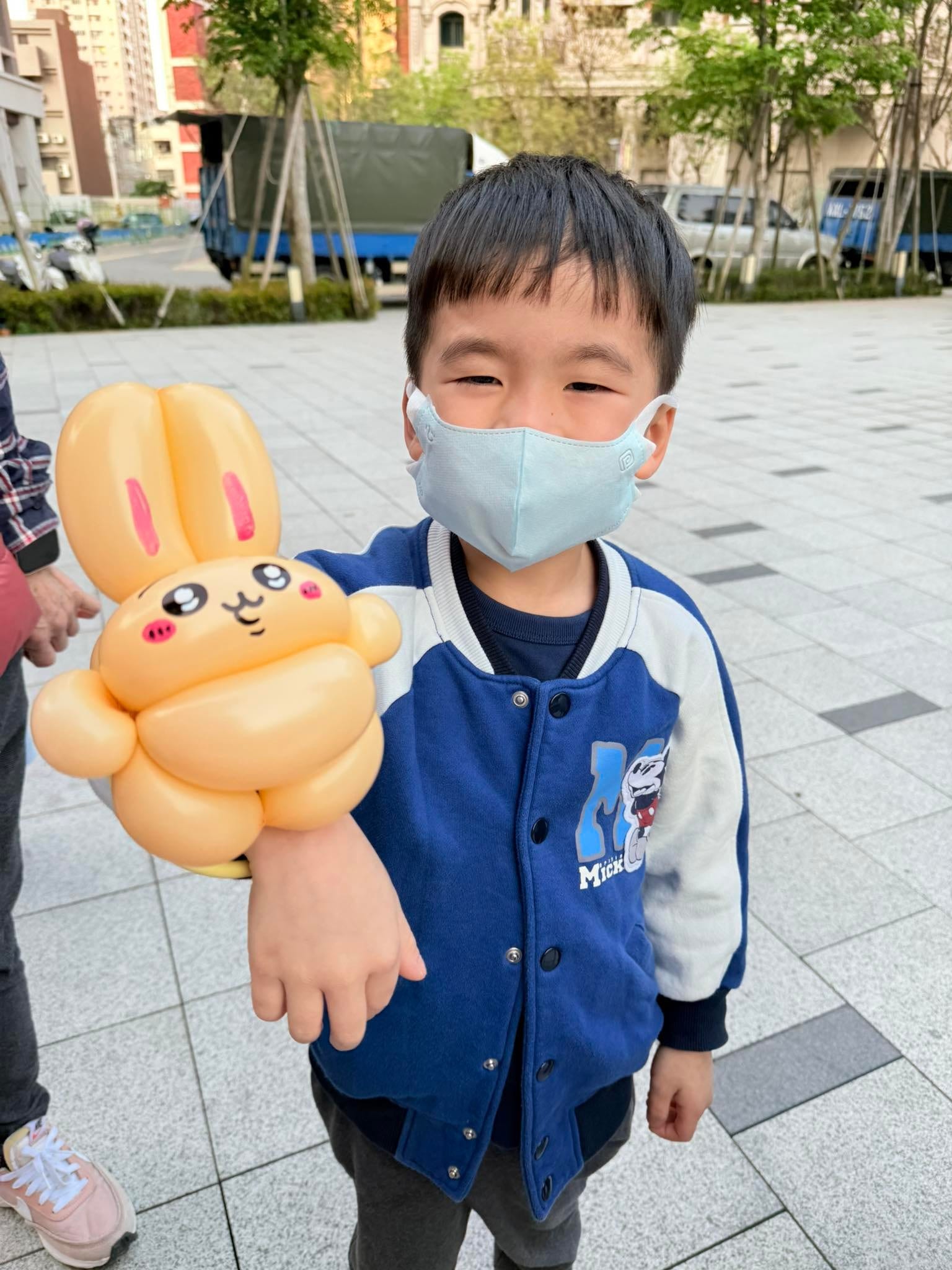 穿藍色棒球外套的男孩手臂套著烏薩奇手環造型氣球，站在市集廣場