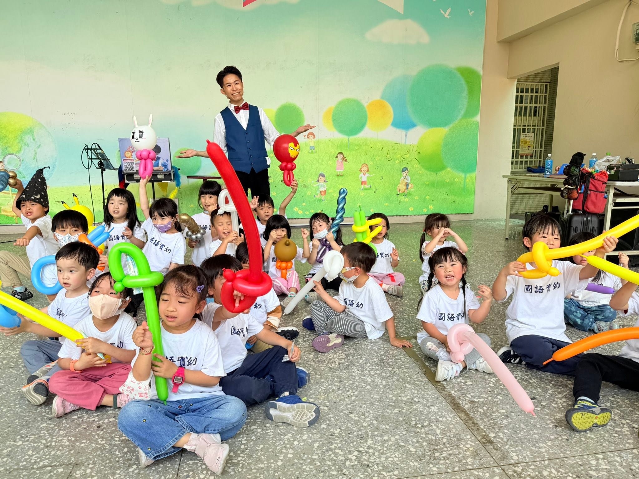 穿國語實幼白色T恤的幼兒園小朋友拿著寶劍、鋼鐵人、白兔造型氣球坐在地上