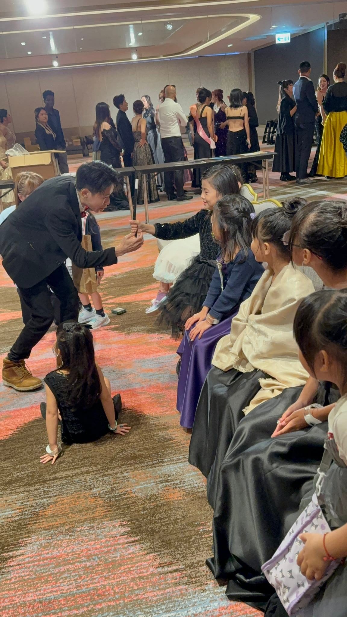 柯男彎腰把黑色魔術棒交給穿黑色紗裙的女孩，旁邊小朋友專心看