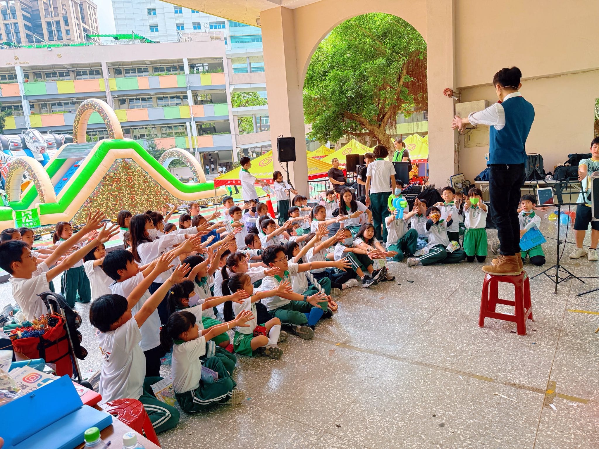 穿綠白制服的小學生坐在地上一起伸手施法，柯男背對鏡頭帶動魔術互動