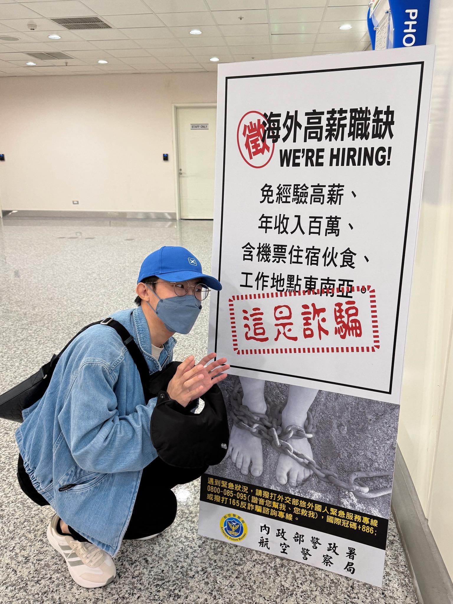 我戴帽子口罩，蹲在機場防詐騙海報旁比手勢