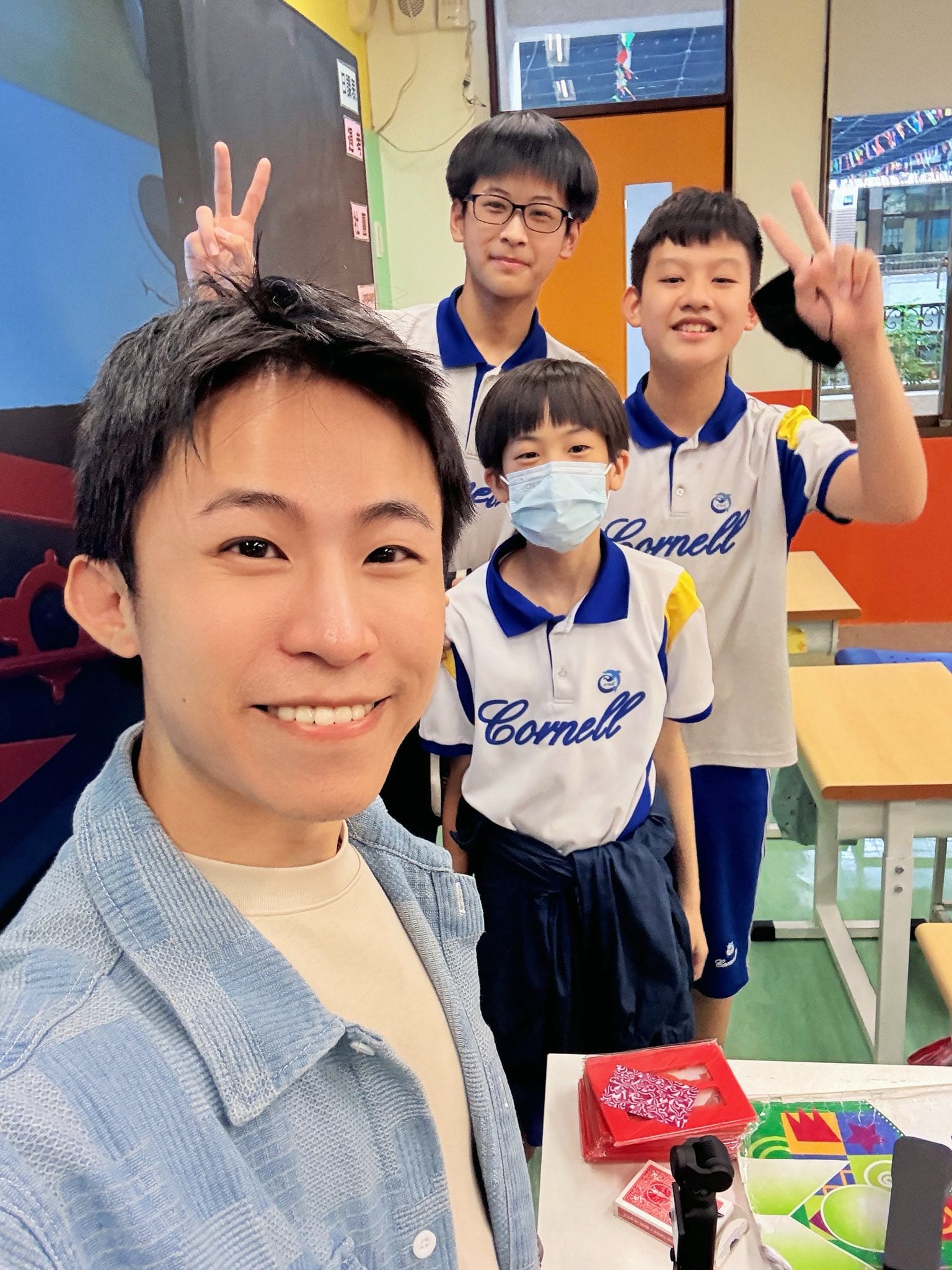 柯男在教室自拍，後方三位比 YA 的男學生，桌上放著撲克牌與魔術道具