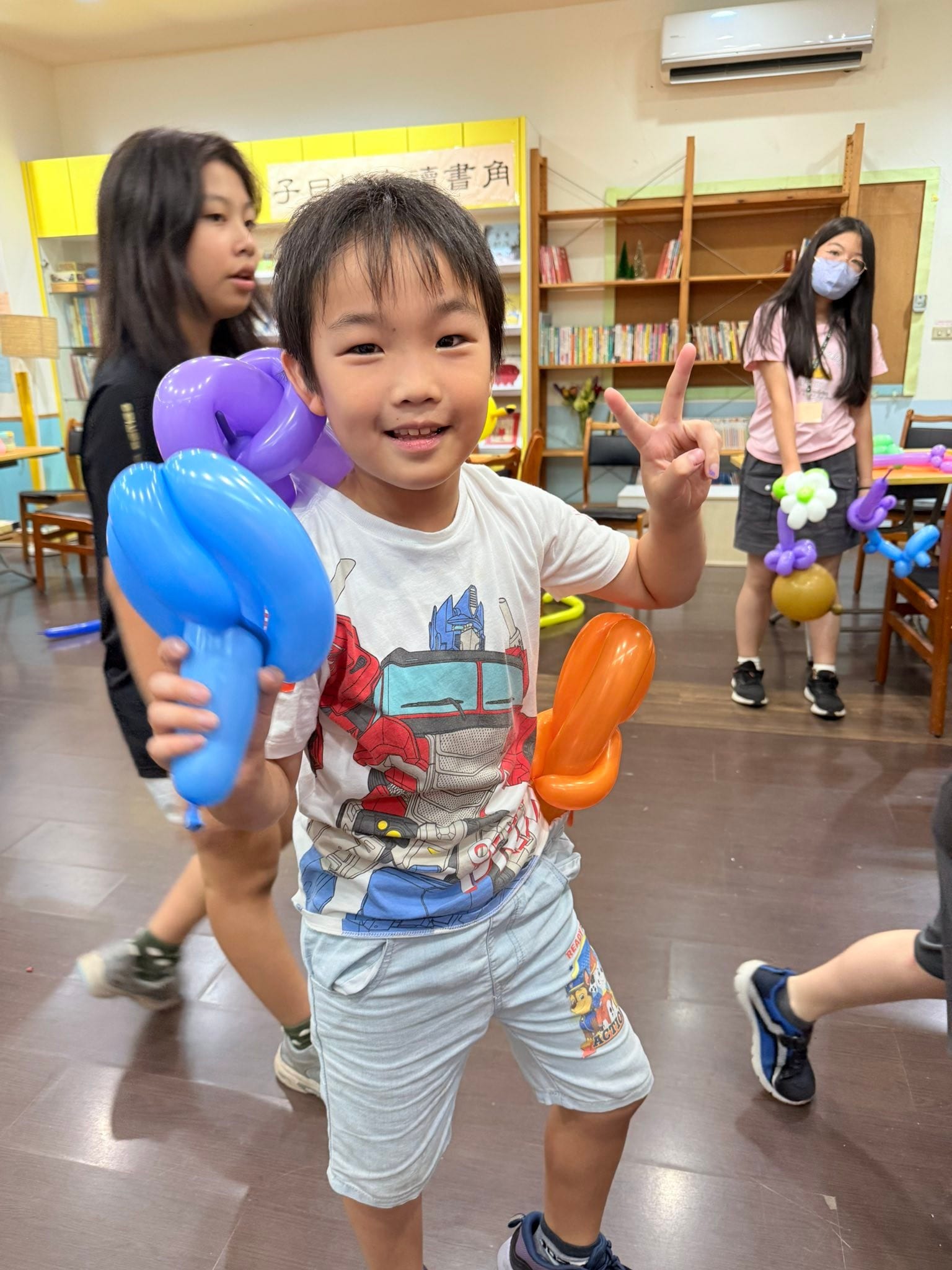 穿白色T恤的男孩比 YA，手上拿著藍色、紫色與橘色的寶劍造型氣球