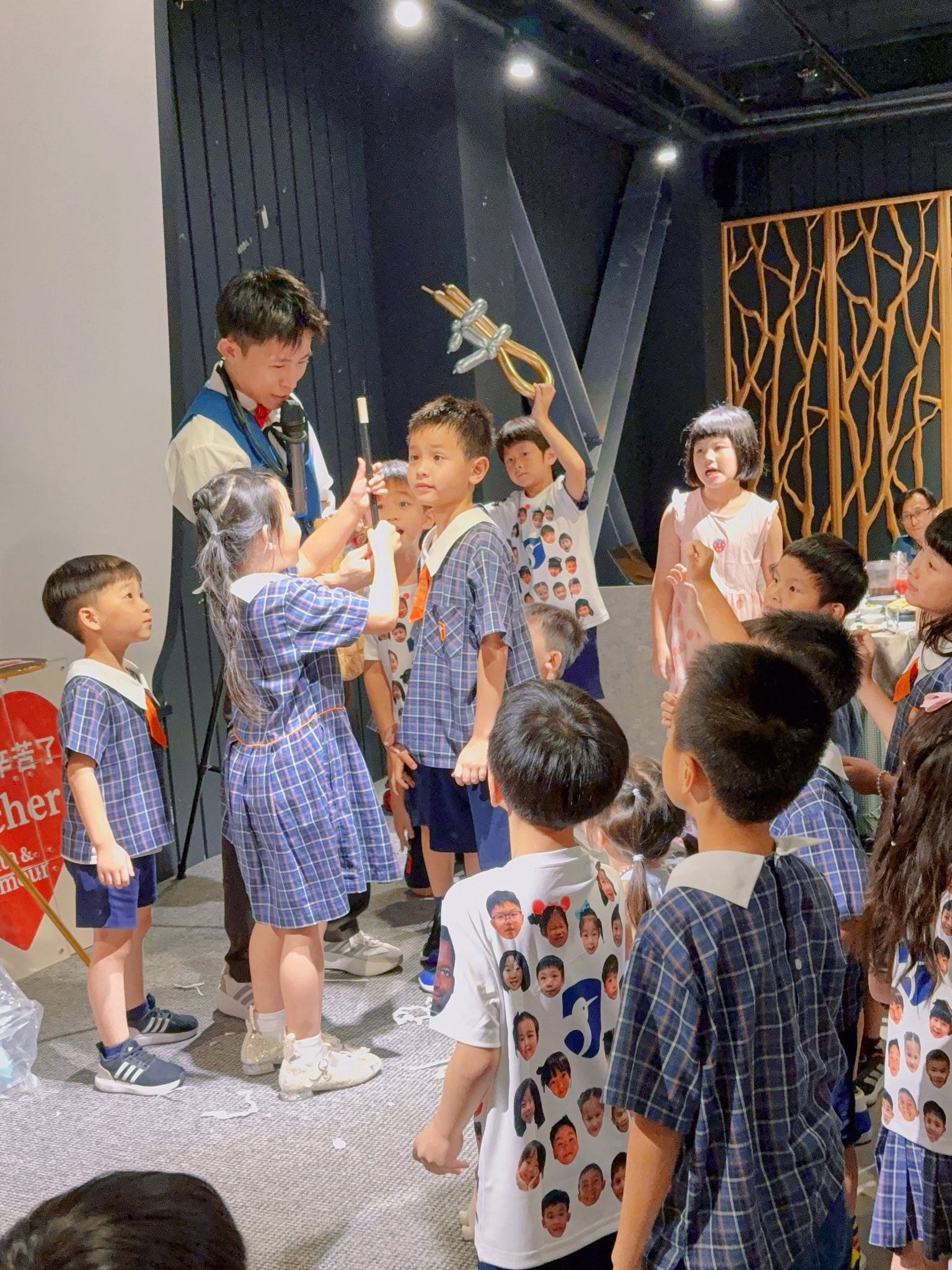 穿格子洋裝的小女孩在台上幫柯男拿黑色魔術棒，旁邊一群小朋友圍在舞台上