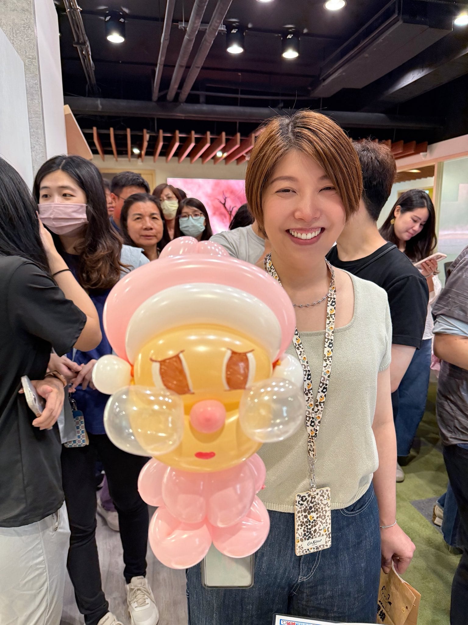 戴識別證的女員工笑著拿起粉紅帽子、大眼睛的娃娃造型氣球，後方同事排隊