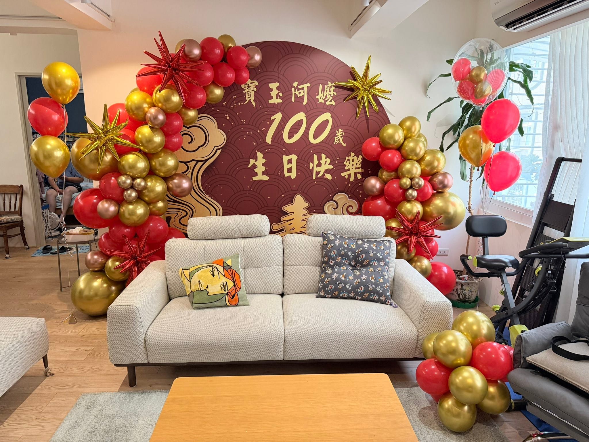 百歲壽宴氣球佈置，客廳沙發後方酒紅色圓形百歲生日快樂背板，兩側紅金氣球拱